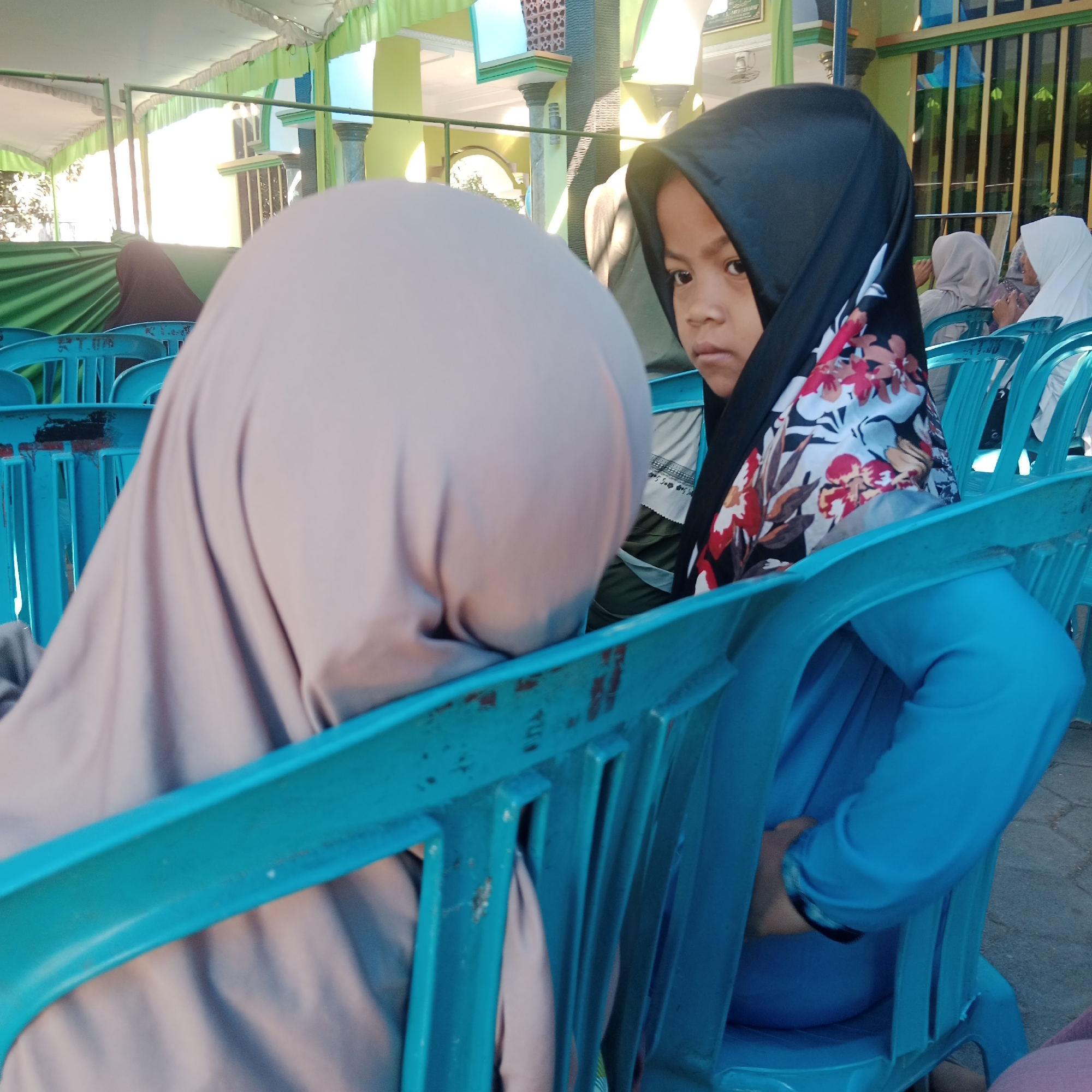 Two young girls, dressed in hijabs, are sitting on blue plastic chairs. The girl on the right is wearing a black hijab with a floral pattern of red and pink flowers. Her face is partially visible, and she is looking forward. The girl on the left is wearing a beige hijab and is facing away from the camera. In the background, other individuals are also seated, and a building with green and white accents is visible. The scene appears to be outdoors, possibly at an event or gathering, in Tegowanu Kulon, Indonesia.
