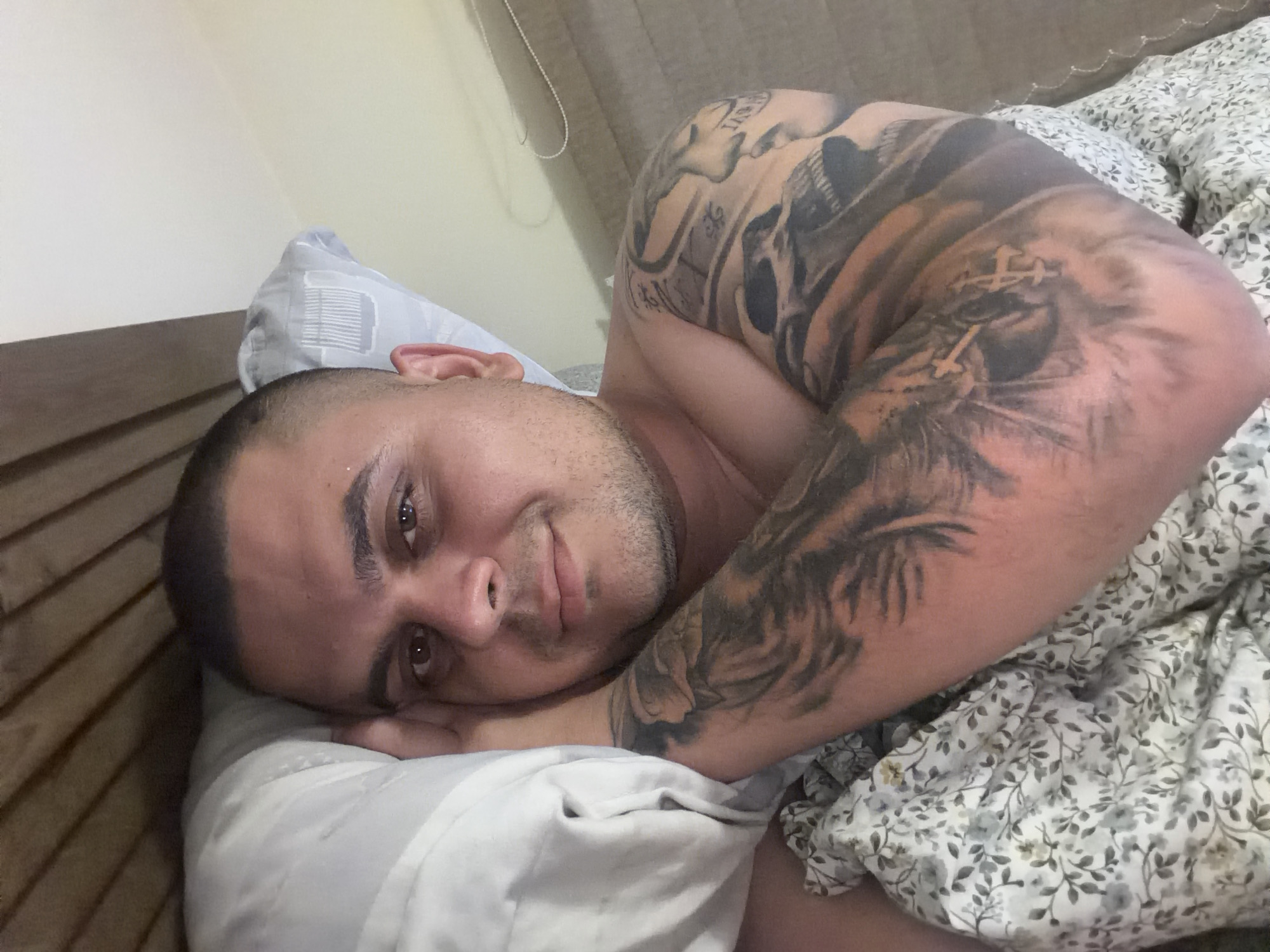 An eye-level, close-up indoor shot captures a man lying on his side in bed, looking directly at the camera with a gentle, relaxed smile. He has a buzz cut and light stubble. His head rests on a light-colored pillow with subtle grey stripe patterns. His left arm, prominently in the foreground, is adorned with an elaborate sleeve tattoo. The tattoo artistry appears to include elements of a skull, a large feline head (possibly a lion or tiger) with what resembles a crown of thorns or crosses above it, and intricate shading and patterns.
The man is partially covered by a light-colored duvet or comforter featuring a delicate floral or leaf pattern in muted grey and light brown/yellow tones. In the background, a wooden slatted headboard is visible to the left, and a plain, light-colored wall occupies the upper area. A textured, light-colored curtain or blind can be seen partially in the upper right. The scene depicts a calm bedroom setting, suggesting a moment of rest or leisure. The location context is Shumen, Bulgaria. No visible text is present in the image.