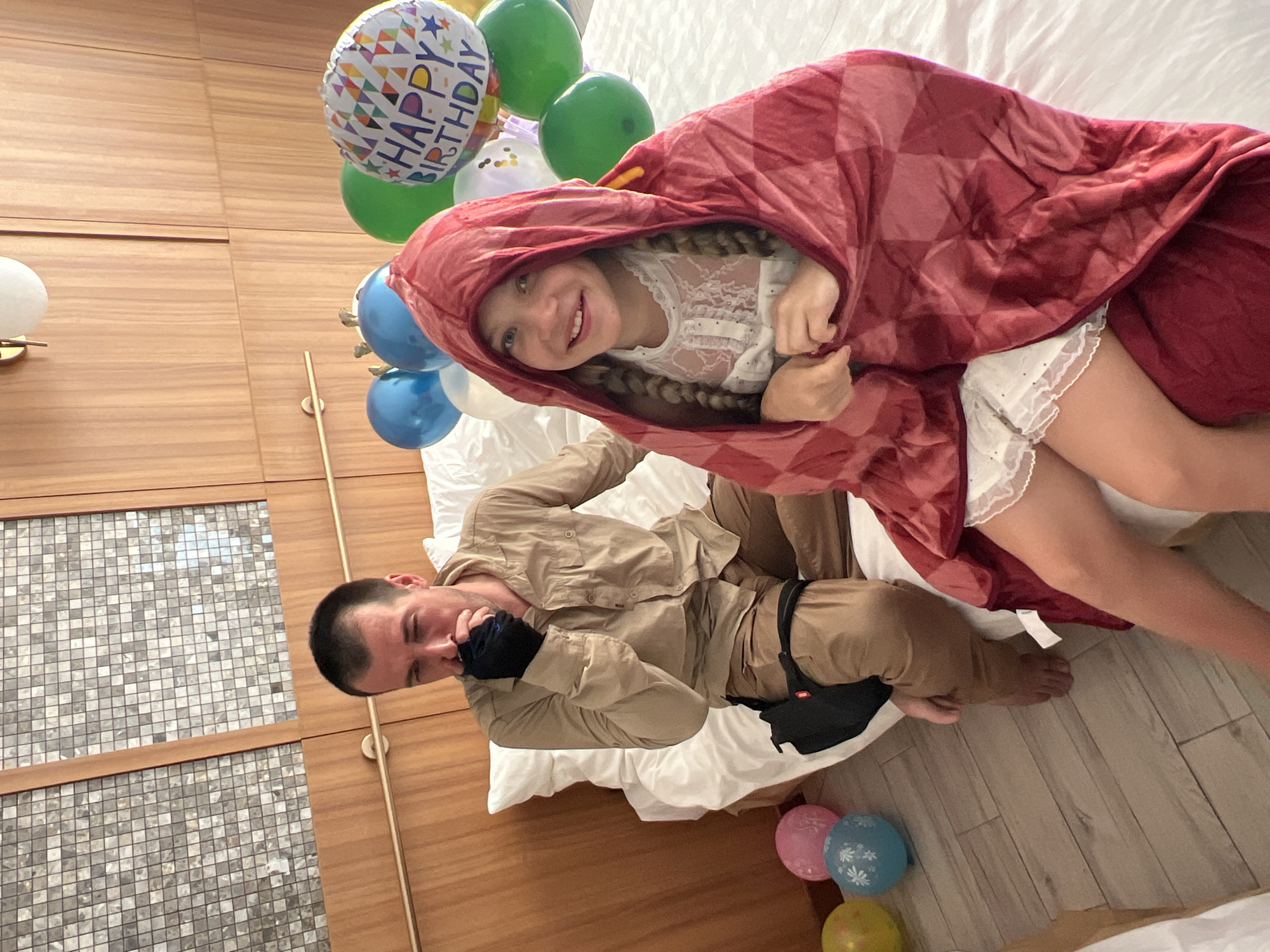 This is a photograph of a young girl and an adult man in a bedroom. The girl is wrapped in a red blanket with a checkered pattern and is peeking out with a smile. She has blonde hair in braids and is wearing a white lace top. The man is lying down next to her, wearing a tan shirt and pants. He appears to be holding his hand to his face, looking somewhat pensive.
Scattered around the bed are several balloons, including one that says "HAPPY BIRTHDAY" in colorful letters. There are also blue, green, white, and pink balloons. The background shows a wooden headboard and a white duvet. The room has a warm, cozy atmosphere, suggesting a celebration or a relaxed moment. The lighting appears to be indoor lighting.
There is no indication of the time of day or the weather outside. The overall scene evokes a feeling of joy and togetherness, likely related to a birthday. The man's expression is somewhat ambiguous, but the presence of balloons and the girl's happy demeanor suggest a positive event.