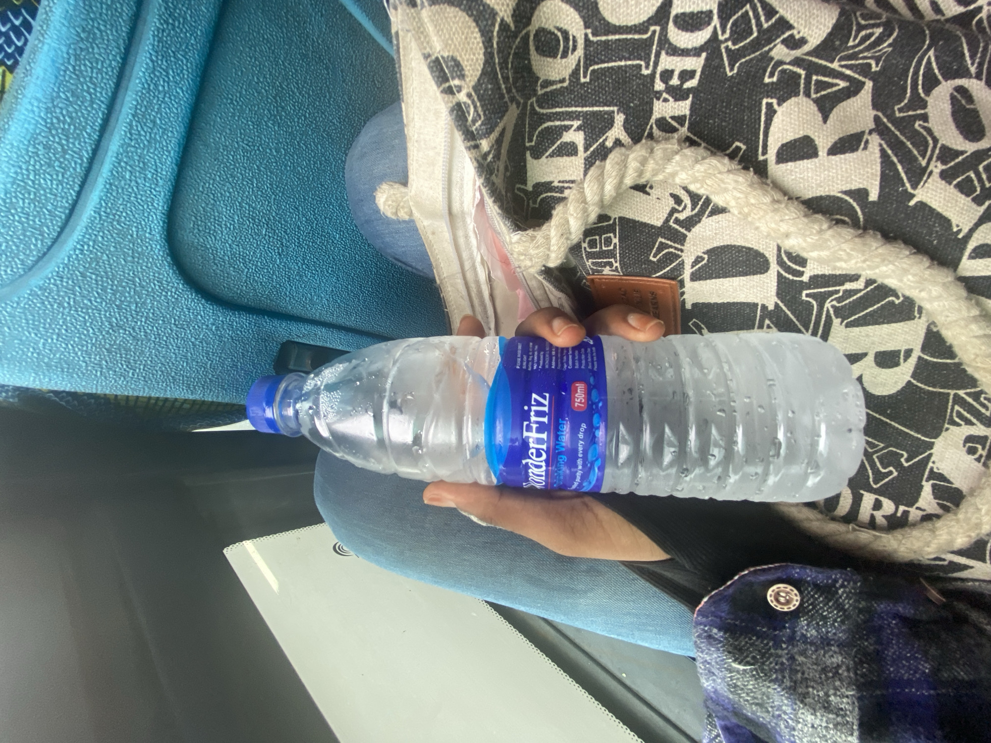 Here is a detailed description of the image:
A top-down shot, likely taken inside a moving vehicle or bus, presents a person holding a clear plastic water bottle. The bottle is labeled "WonderFriz" and contains condensation, suggesting it's cold. The person's hand is visible holding the bottle. The person is wearing blue jeans, a black top, and a blue-checkered shirt with a button visible.
The water bottle and hand rest on the person's lap, with part of a patterned tote bag in the background. The bag has white rope handles. The interior of the vehicle is visible, including part of a blue seat and a window with a white card or paper attached.