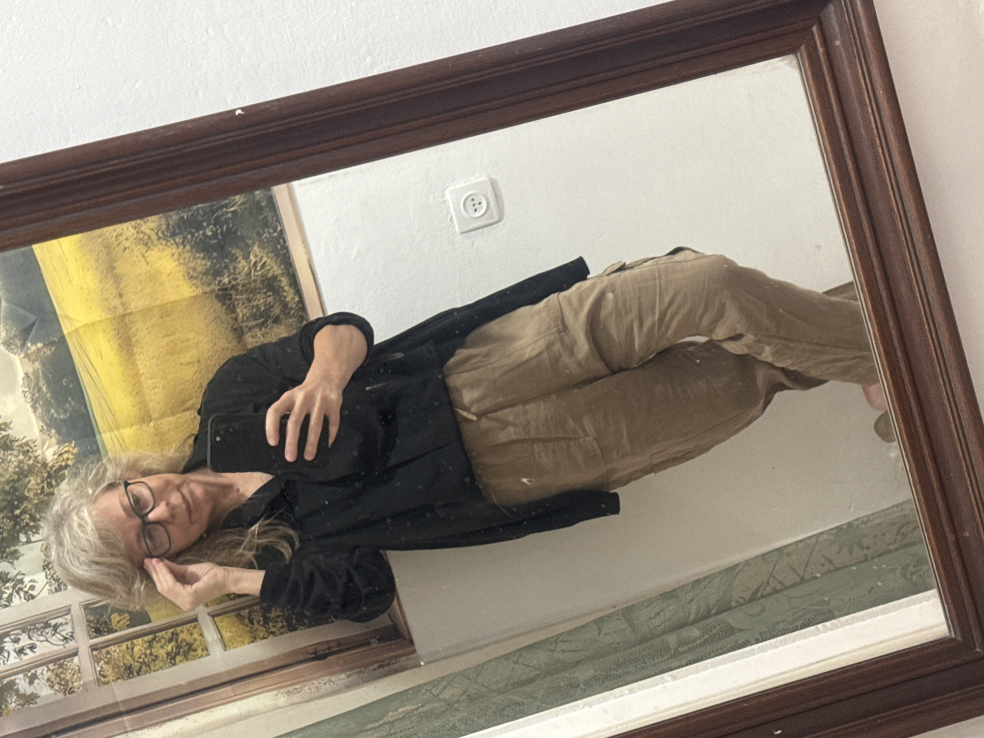 The image appears to be a selfie taken in a mirror. The individual in the mirror is a woman with gray hair and glasses, wearing a black shirt and khaki pants. She is holding a smartphone. Reflected in the mirror are a painting or print, a light switch, and what seems to be wallpaper or a decorative border on the wall. There is no information provided in the image to suggest it was taken in Ramat Gan, Israel. The location is undetermined based on the visible elements.