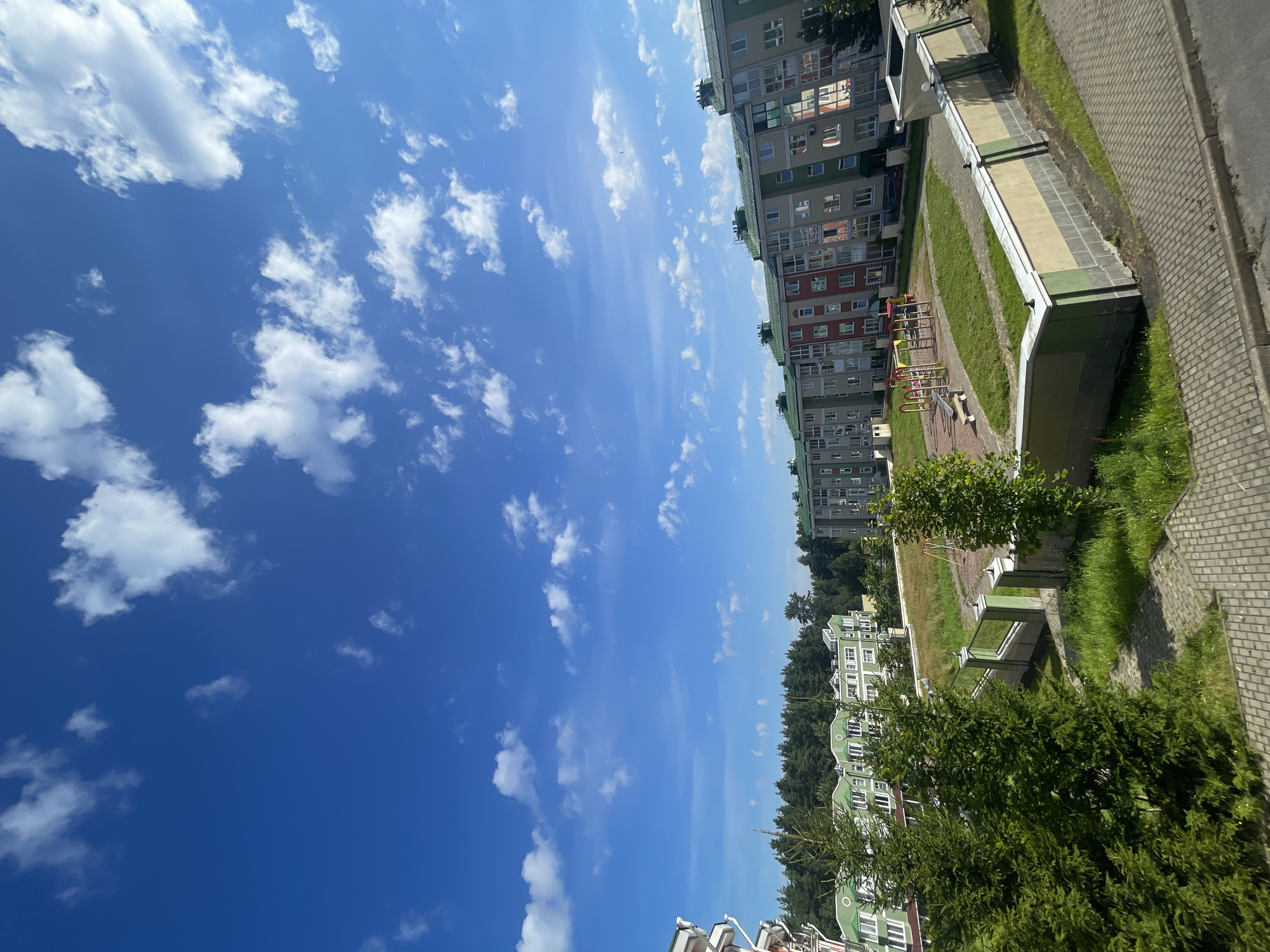 The image depicts a clear blue sky with scattered white clouds over a residential area in the city of Kaltino, Russia. In the foreground, a paved walkway with a curb borders a grassy area. A low wall, partially concrete and partially plastered in yellow and green, surrounds a playground. The playground features colorful swings, slides, and a bench. Beyond the playground, several multi-story apartment buildings are visible. Some of these buildings have green roofs, and their facades are a mix of light gray, beige, and red. Behind the apartment buildings, a dense line of evergreen trees forms a forest. The overall scene suggests a suburban or semi-urban environment on a sunny day.