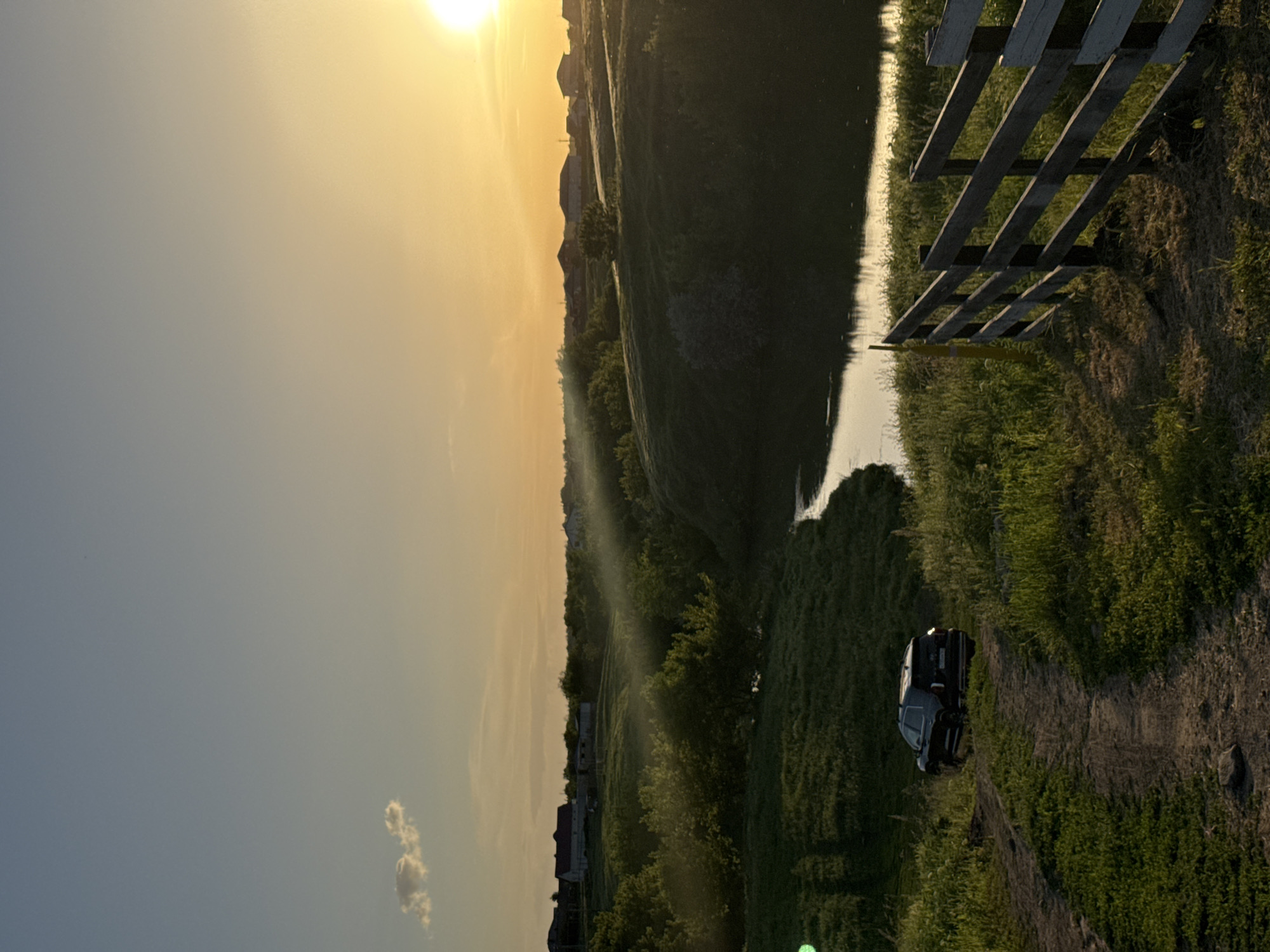 The image depicts a rural scene at sunset, likely taken in Усть-Курдюм, Russia. The setting sun casts a golden glow across the sky, with a few scattered clouds visible. The landscape features a body of water, potentially a river or stream, bordered by grassy banks. A partially obscured wooden fence runs along one side of the waterway. A dark-colored vehicle is parked on a dirt path near the edge of the grass. The environment consists of overgrown grass, trees, and possibly some residential buildings further in the background. There are no people visible in the immediate scene.
