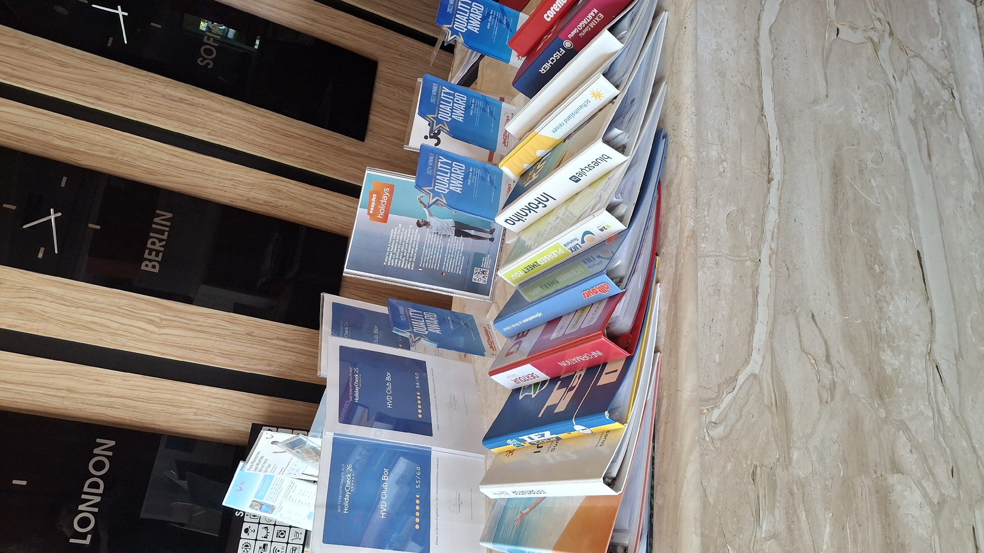 This image depicts a display of brochures and binders on a marble counter, possibly in a reception area or promotional space. Behind the display, a wall features wooden slats and black panels with city names and clock faces, indicating different time zones. The city names visible are "LONDON," "BERLIN," and "SOFIA." The brochures and binders appear to be related to travel and holidays, with titles such as "HolidayCheck," "HVD Club Bor," "QUALITY AWARD," "easyjet holidays," and "InfoKinaha." Some brochures are displayed in clear plastic stands. The binders are arranged in a row, with their spines facing outwards, showcasing various colors and text. The lighting suggests an indoor environment.