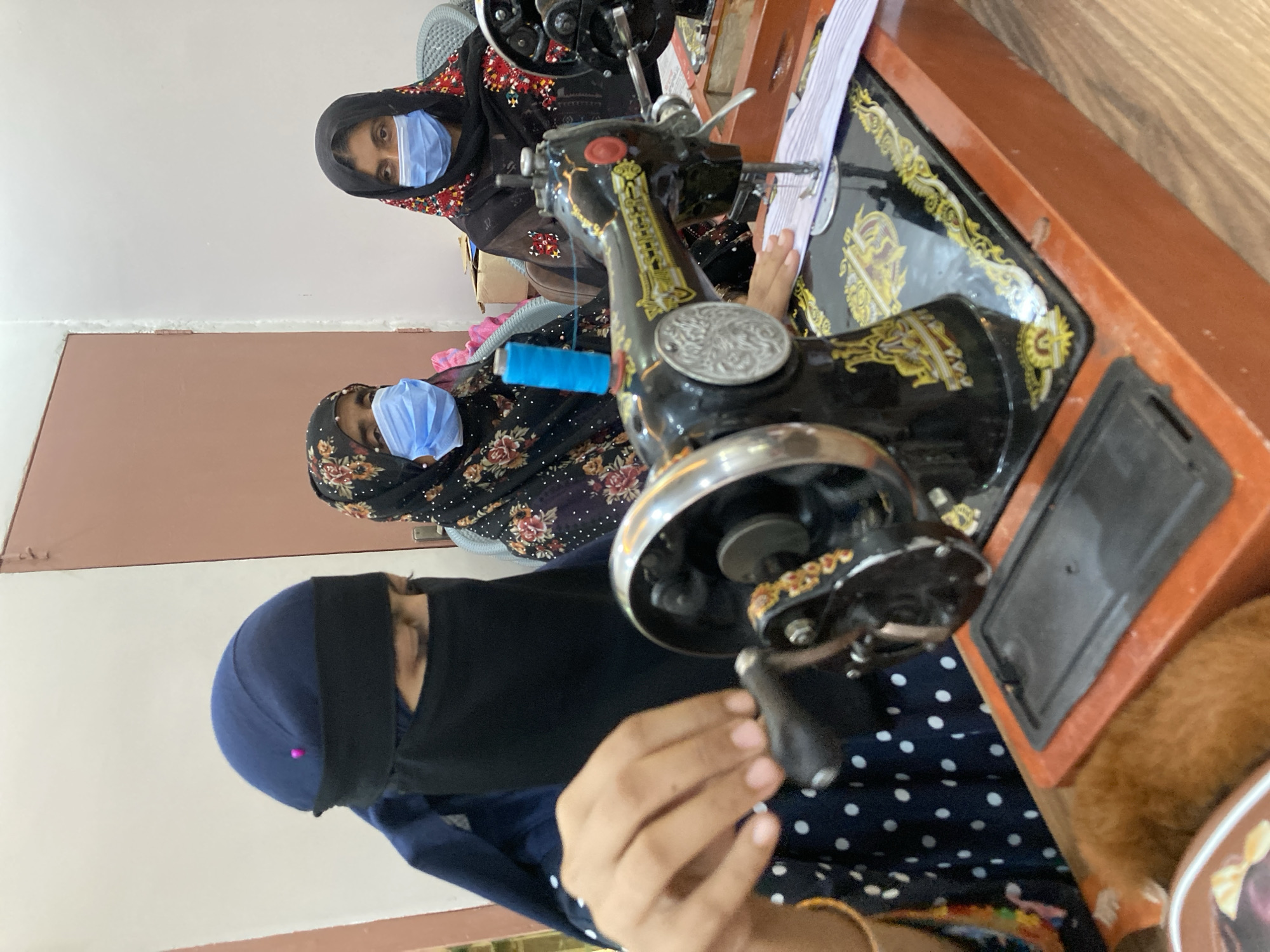 Three women wearing face masks and head coverings are operating sewing machines. The women are positioned around the sewing machines, focusing on their work. The sewing machines are vintage models with black and gold detailing. The environment appears to be an indoor setting, possibly a workshop or studio, located in Rāwāni, Pakistan. The overall scene depicts a work environment with a focus on sewing.