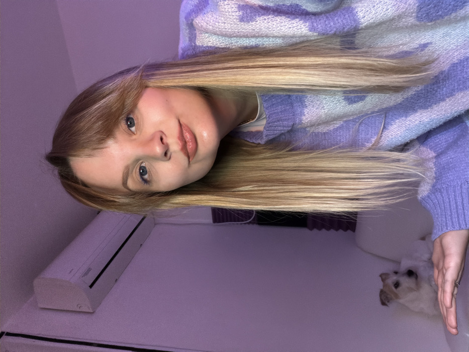 An indoor portrait features a young woman with long, straight blonde hair and bangs, looking directly at the camera with a slight smile. She has light skin and blue eyes, and her lips appear glossy. She is wearing a soft, chunky knit sweater with a striped or patterned design in shades of light purple, lavender, and white. Her hair flows down past her shoulders.
To the left of the woman, mounted on a light-colored wall, is a white rectangular air conditioning unit. The room's walls have a prominent light purple hue, which might be due to ambient lighting or the wall color itself. In the bottom right corner of the frame, a small dog with white and light brown fur is partially visible, lying down or looking up towards the woman. Behind the dog, part of a white or light-colored bed or sofa is visible. The overall setting is a casual indoor space, possibly a bedroom or living room in Tel-Aviv, Israel, as per the provided context. The lighting suggests an indoor, possibly evening or nighttime, environment. There is no visible text in the image.