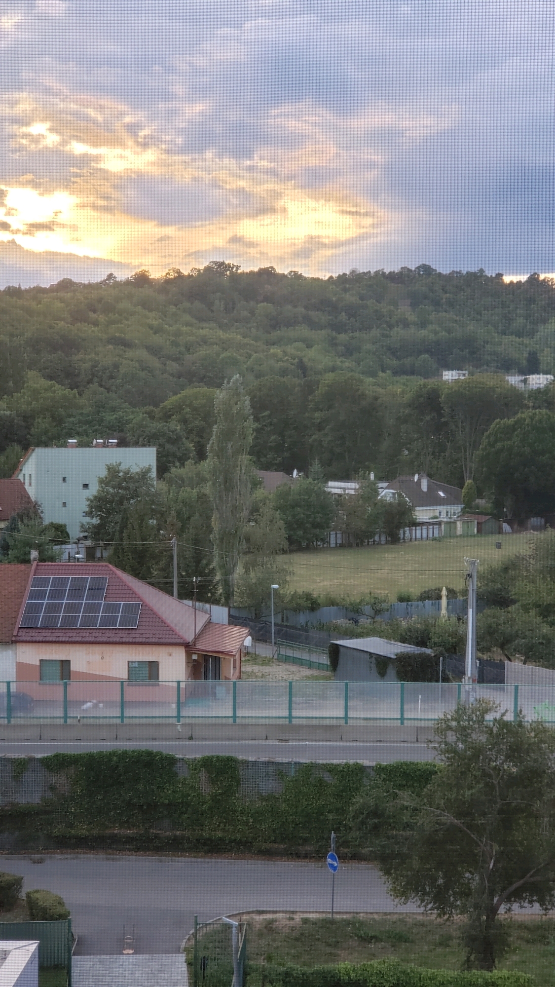 The image depicts a suburban landscape with a densely wooded hill in the background. The sky is partly cloudy with the sun setting, casting a warm glow on the clouds. In the mid-ground, there are several residential buildings, including a house with solar panels on its roof. A road runs horizontally across the lower half of the image, bordered by greenery. A blue traffic sign indicating a speed limit is visible on the right side of the road. The scene appears to be taken from a window with a screen, which creates a grid-like overlay on the image. The overall impression is one of a peaceful, late afternoon in a residential area.