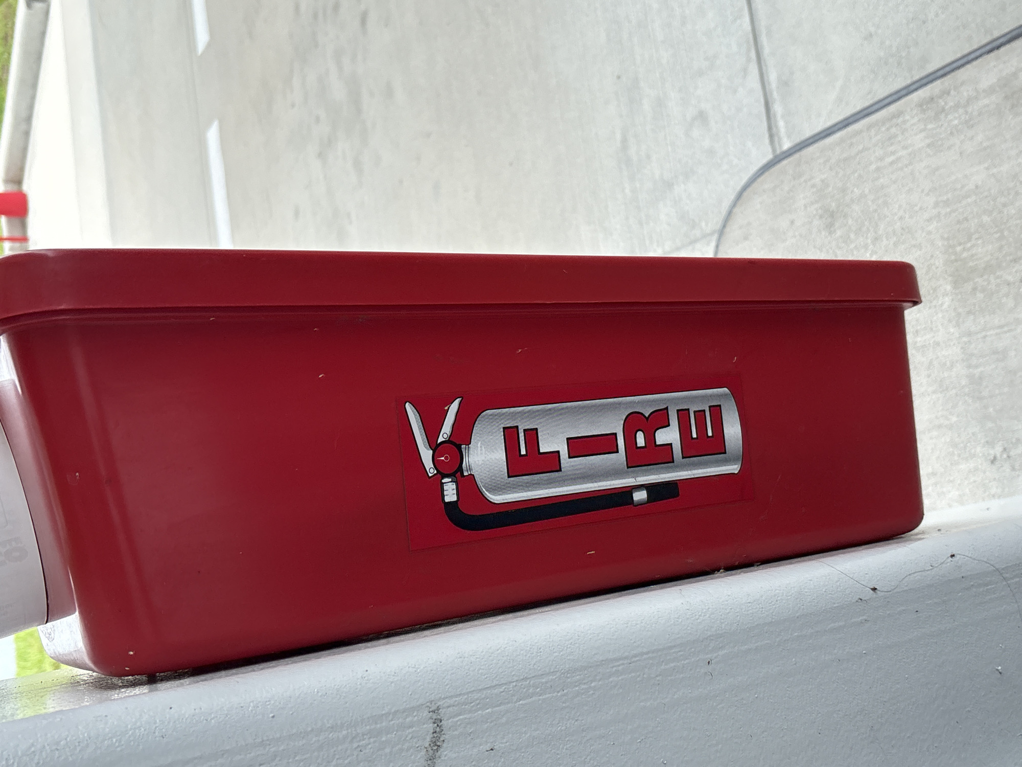A close-up, vertical, eye-level shot shows a red fire extinguisher case mounted on a white pole. A sticker of a fire extinguisher with the word "FIRE" in red lettering on a silver background is affixed to the red case. The case is a vibrant red, and the white pole is to the left of the case, appearing to be cylindrical or rectangular. The background is blurred, showing a gray concrete surface with white lines painted on it, suggesting an outdoor area like a parking lot or sidewalk. Some greenery is visible in the far background. The lighting appears to be natural daylight, casting subtle shadows. The image is sharp and detailed, focusing on the fire extinguisher case and its sticker.