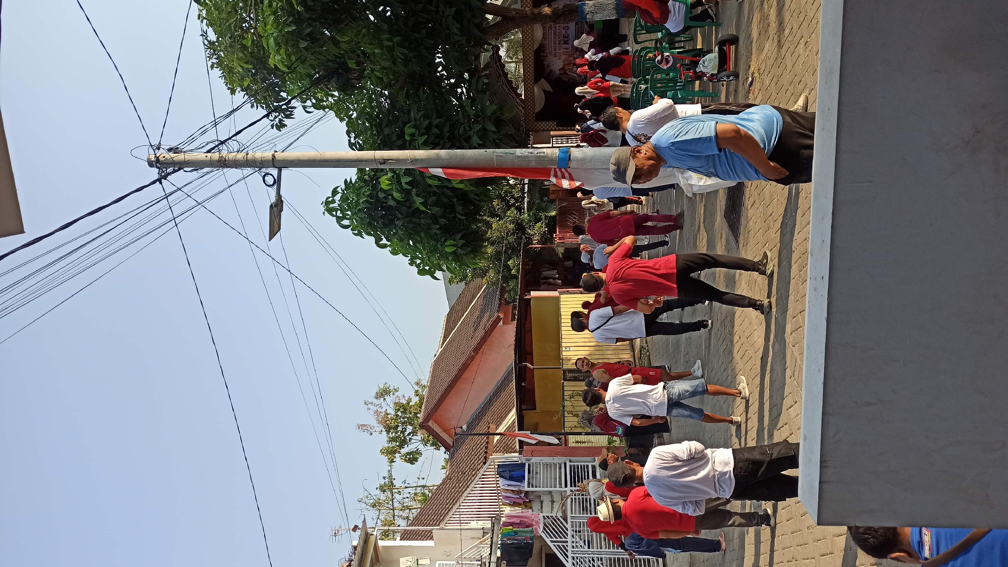 In Galak, Indonesia, a group of people are gathered outdoors, likely for a community event or physical activity. The scene is set in a paved area, possibly a public square or street, with houses visible in the background. The sky is clear and blue, suggesting a sunny day.
Several individuals are actively engaged in what appears to be exercise or a group dance, their postures indicating movement and coordination. Many are dressed in casual attire, with a prominent color scheme of red and white evident among some participants, possibly signifying national pride or a specific event. A few people are seated on green chairs, observing the activity.
Objects in the environment include a utility pole with numerous wires, a street lamp attached to it, and trees providing shade. A small red and white flag is displayed on a pole near the houses. Laundry is hanging on a rack outside one of the houses. The overall atmosphere seems to be one of communal gathering and shared activity in a residential area.