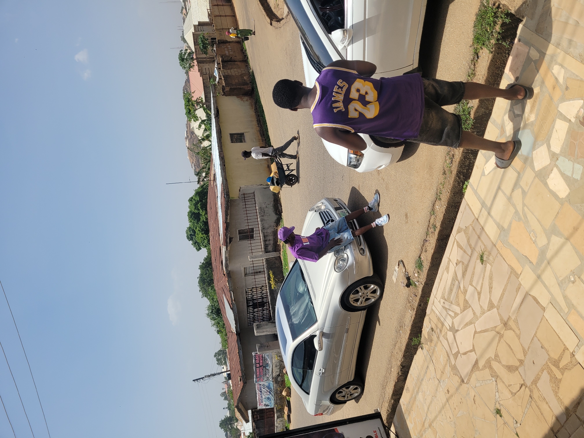 The image shows an outdoor scene in Jos, Nigeria. A white Mercedes-Benz car is parked on a narrow road. A young person wearing a purple shirt and a matching hat is sitting on the hood of the car. Another young person, wearing a purple and yellow jersey with the number "23" and the name "JAMES" on the back, is standing next to another white car. A person is walking with a bicycle on the side of the road. Houses and buildings are visible in the background. The sky is clear and blue. The ground is paved. The image shows a poster for the "Home of Grace Gospel Ministry Academy" and a table.