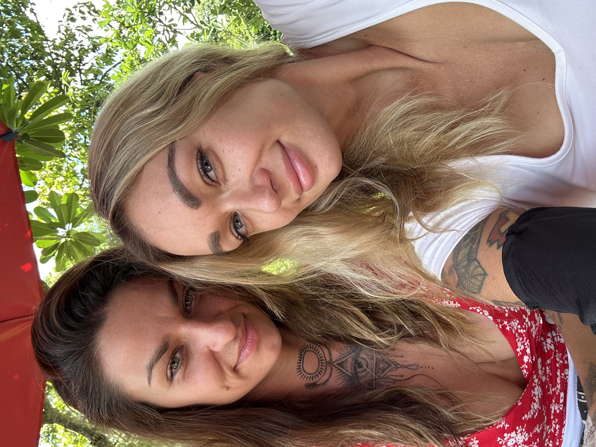 Two women are posing for a selfie, possibly outdoors in Dĩ An, Vietnam. The woman at the top has blonde hair and is wearing a white top. The woman at the bottom has brown hair and is wearing a red and white floral-patterned dress. She also has tattoos on her neck and arm. Green foliage and an umbrella are visible in the background.