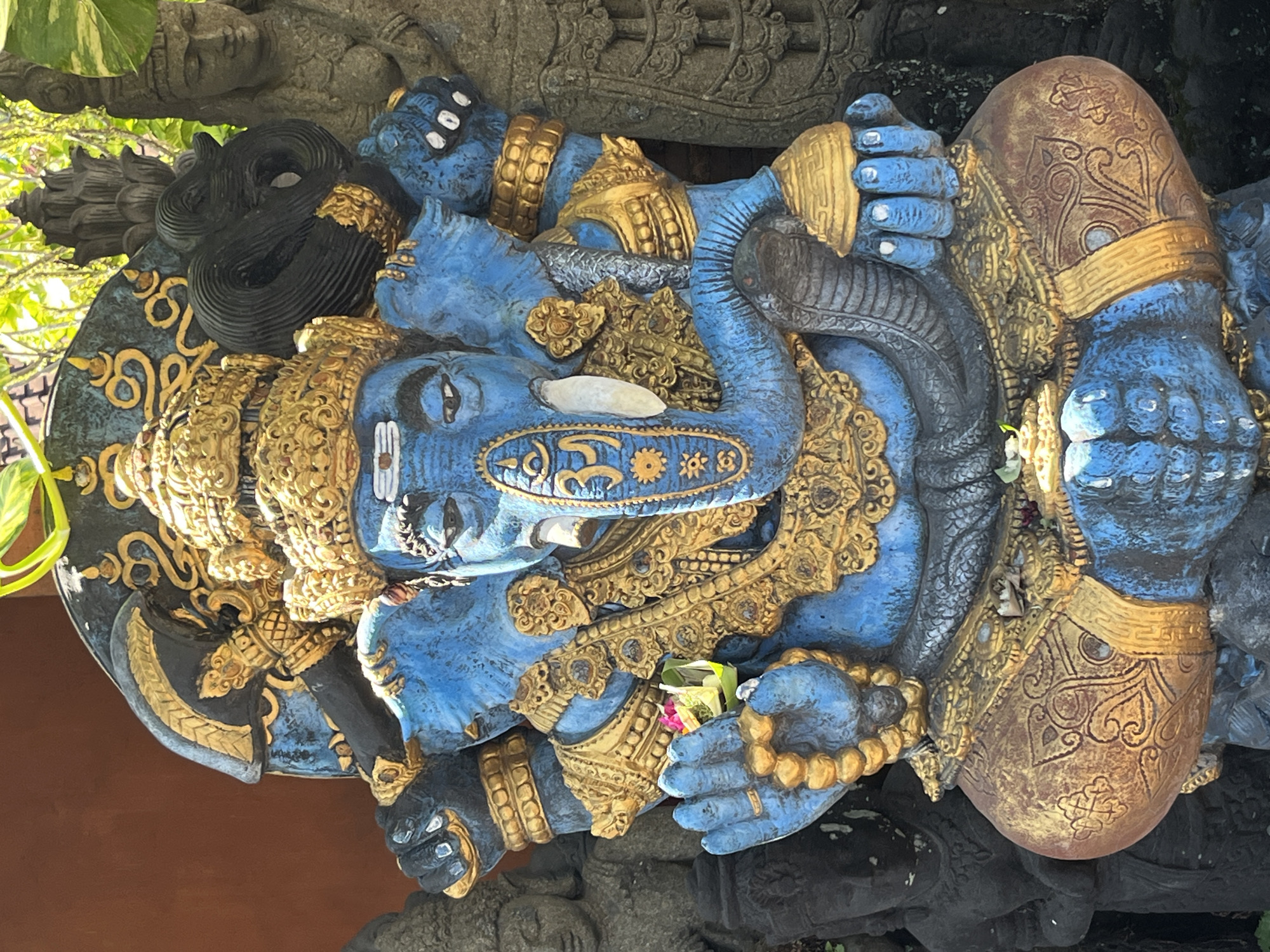 A vibrant blue statue of Ganesha, a deity with an elephant head, sits in a cross-legged position. The statue is adorned with intricate gold ornamentation, including a crown, jewelry, and decorative patterns on its robes. Ganesha's trunk is curled and rests on his chest, and one of his hands holds a small offering of flowers. The statue is positioned in an outdoor setting, with a reddish-brown wall visible in the background and lush green foliage peeking from the top and right sides. Other stone statues, appearing to be part of a garden or religious site, are partially visible on the left and right edges of the frame. The overall impression is one of a sacred and artistic display. This statue is located in Sukawati, Indonesia.