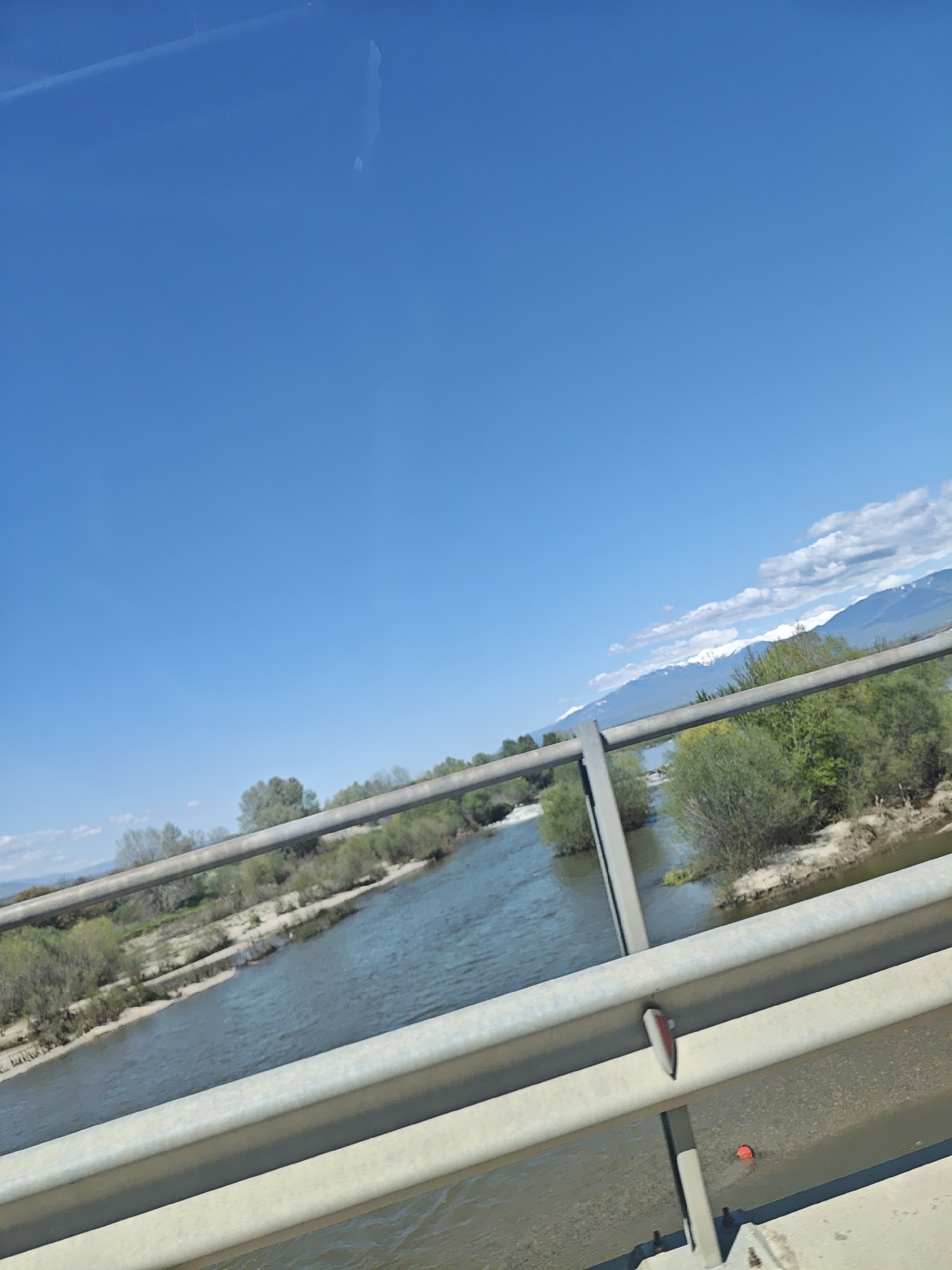 The image shows a river flowing under a bridge, as viewed from the bridge. The bridge has gray metal guardrails. The river is flanked by sandy banks with some vegetation. In the distance are mountains with some snow on their peaks and clouds in the sky. The sky is a clear blue. The location is in an unknown city.