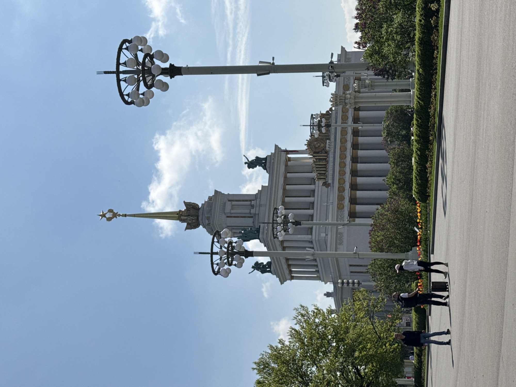 The image captures a view of the Central Pavilion at the Exhibition of Achievements of National Economy (VDNKh) in Moscow, Russia. The architectural details of the pavilion, including its columns, statues, and the spire topped with a star, are prominent. Streetlights with circular clusters of globes are also visible. Three people are walking on the pedestrian pathway to the right of the building. Trees and shrubs are interspersed around the building. The sky is blue with scattered white clouds.