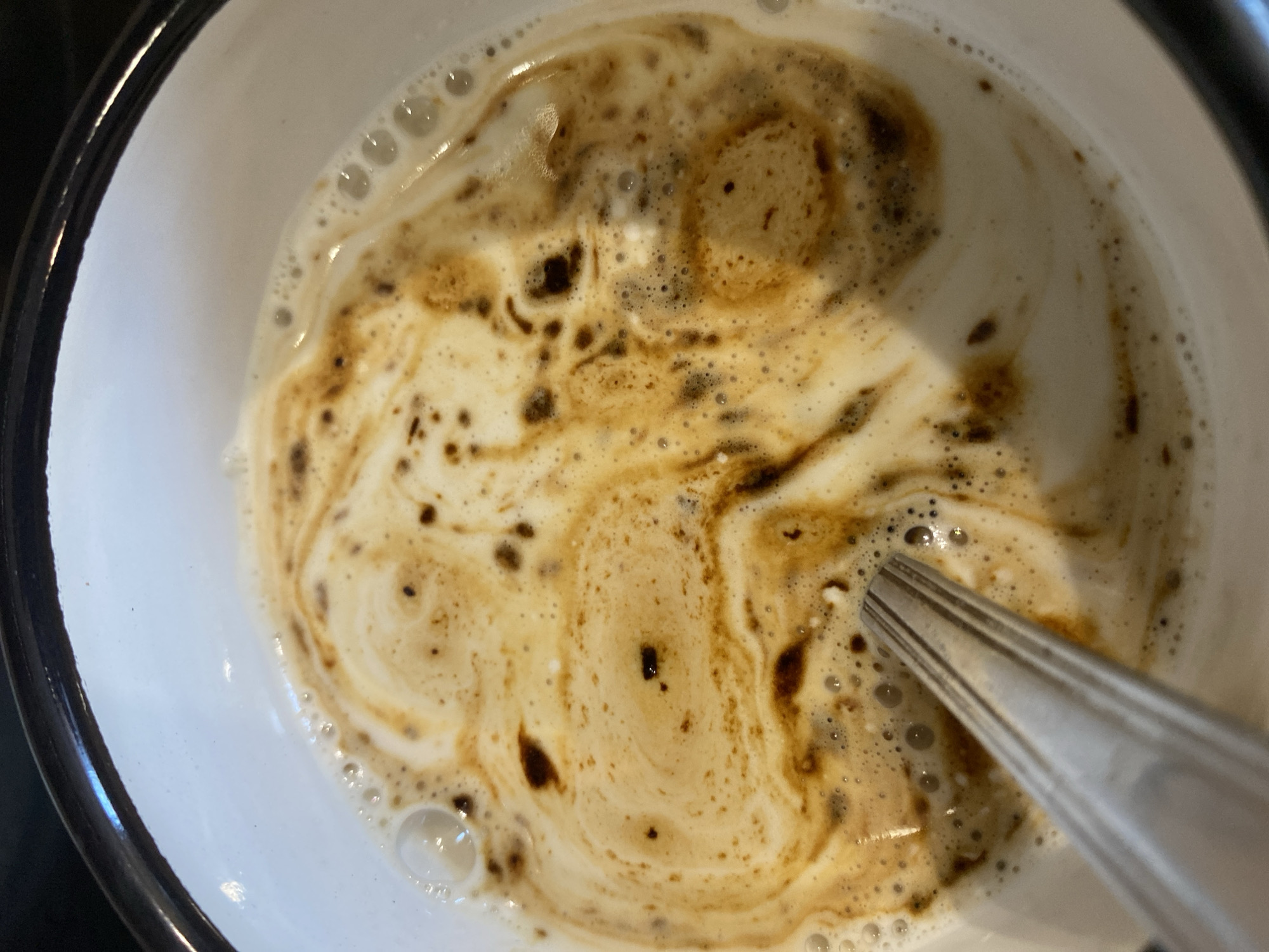 A close-up, overhead shot shows a white bowl with a dark rim filled with a light-colored liquid, possibly milk or cream, swirled with darker brown liquid, likely coffee or chocolate. Small bubbles form a frothy layer on the surface. A silver spoon with a ribbed handle is submerged in the liquid, angled from the bottom left towards the center. The swirls in the liquid create an abstract pattern. There is no indication of the city of Tyumen, Russia, within the image.