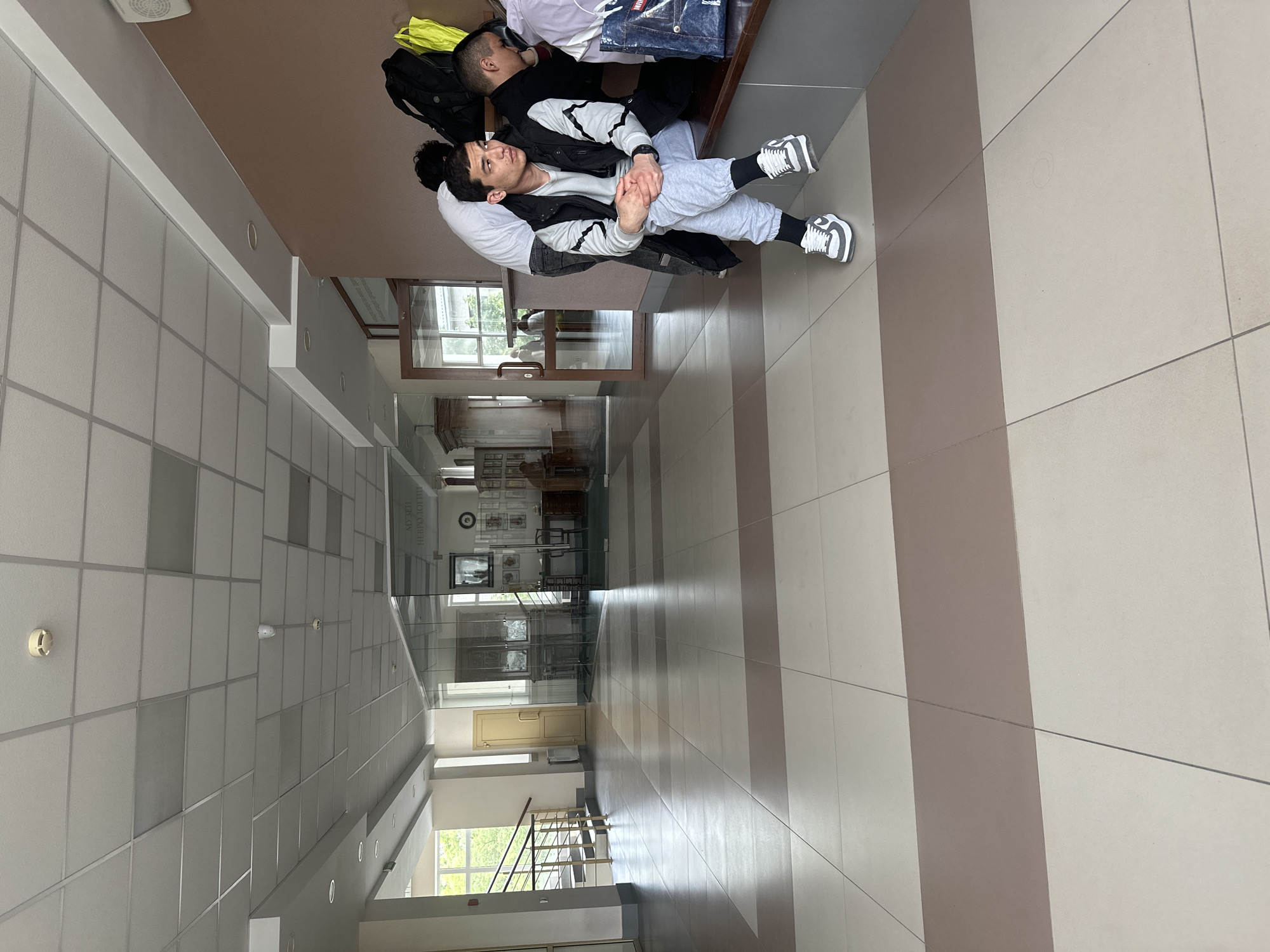 Three men are resting on a wooden bench in a bright, modern indoor hallway in Kazan, Russia, likely within a medical or educational institution housing a "Museum of Neurology." The image, originally rotated, depicts a long corridor with a distinctive floor pattern of alternating light beige and brown rectangular tiles.
The foreground features one man seated on the bench, facing the camera with his hands clasped. He wears a black and white hooded jacket, light grey sweatpants, and white sneakers with black accents. Behind him, another man leans back on the bench, partially visible, wearing a white t-shirt and possibly a black backpack. A third man is partially visible to the right. On the bench next to them are a black backpack, a bright yellow/lime green bag, and some folded clothing, including a blue denim item with "DENIM" visible text.
The hallway is characterized by a white false ceiling with square tiles and recessed lighting. The walls feature a lighter tone, with glass partitions separating the corridor from adjacent rooms or display areas. Through these glass partitions, an office-like setting and a visible sign reading "МУЗЕЙ НЕВРОЛОГИИ" (Museum of Neurology) are seen, along with display cases possibly containing skeletal or anatomical models. A round clock is also visible on a wall in the background. Further down the hall, a light-colored door and a set of stairs with metal railings can be observed through another glass section. Windows along the side of the building allow natural light into the hallway, revealing green foliage outside, indicating it is daytime. The overall atmosphere suggests a public building, possibly a university or hospital, where the men are waiting or taking a break.
