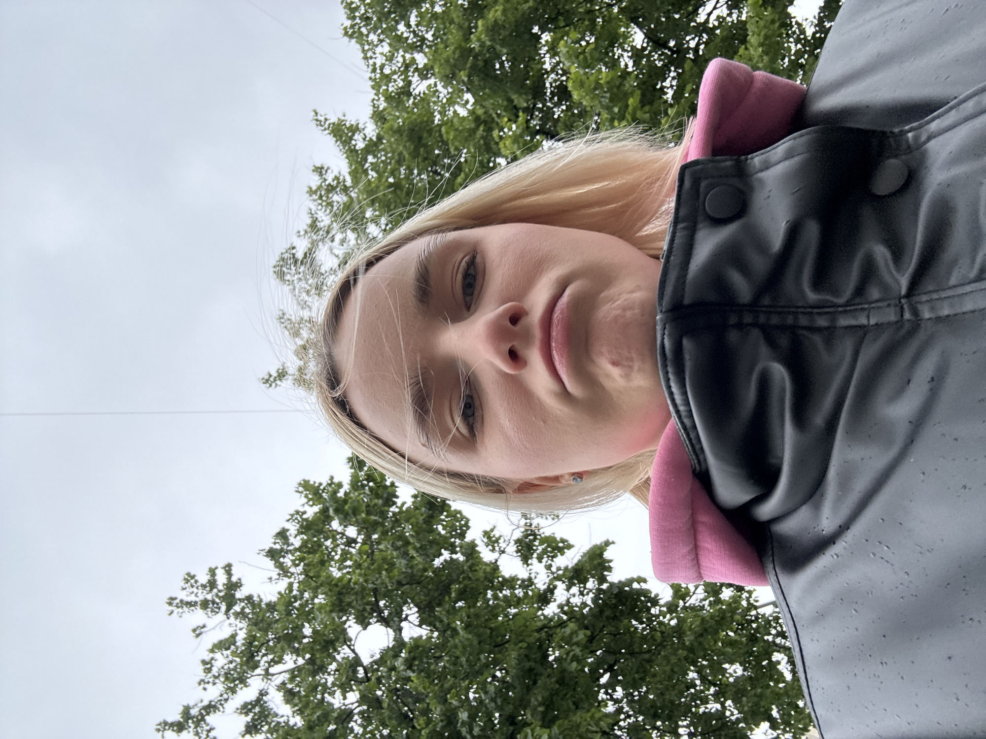 A woman with blonde hair is looking down at the camera. She is wearing a black jacket and a pink hoodie. The background consists of green trees and a gray sky. The location is likely outdoors in an urban environment, as indicated by the trees and the possibility of the sky being visible through buildings in the city of Unknown, Russia.