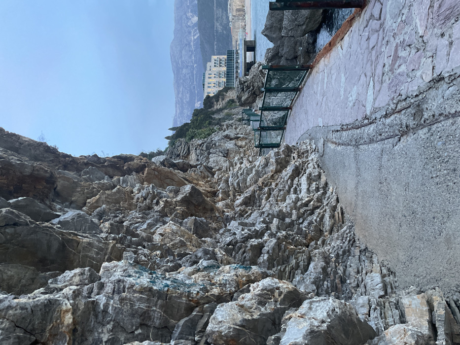 The image displays a coastal scene in Budva, Montenegro. A massive, stratified grey rock cliff, exhibiting distinct layers and varying textures with a patch of turquoise discoloration, occupies the left foreground and mid-ground. At the base of the cliff, a paved pathway curves towards the right and recedes into the distance. This pathway consists of a narrower, lighter grey concrete section adjacent to the cliff, and a wider section of irregular reddish-grey flagstones along its outer edge. A dark green metal fence with vertical bars runs along the right side of the pathway, serving as a barrier to the sea. Dark blue-grey seawater is visible beyond the fence, with several jagged rock formations protruding from it. In the mid-distance, a multi-story building with a light-colored facade and multiple arched windows is situated on the coastline, featuring a concrete structure that extends into the water. Further along the coast, a stretch of sandy beach is discernible. Hazy, grey mountains rise in the far background beneath a pale grey-blue sky. No individuals, animals, or active subjects are present in the frame.