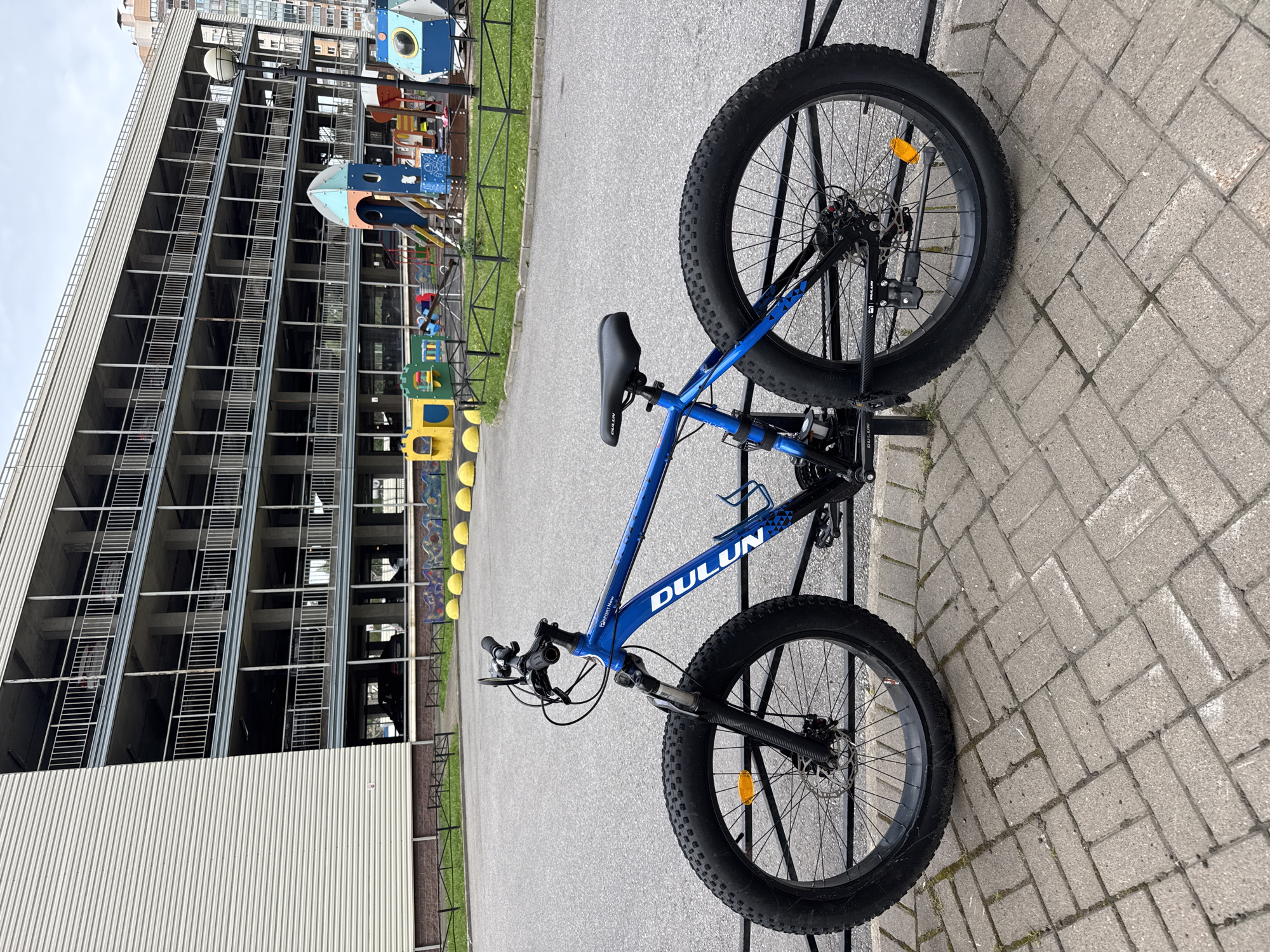 In the city of Sinyavino, Russia, a blue fat-tire bicycle is parked outdoors on a paved walkway. The bicycle, with its wide black tires and black seat, is positioned in front of a black metal fence. Behind the fence, a playground with colorful structures, including a rocket-shaped slide and yellow spherical bollards, is visible. Further in the background, a large, multi-story parking garage with concrete columns and light-colored siding dominates the scene. The sky is overcast.