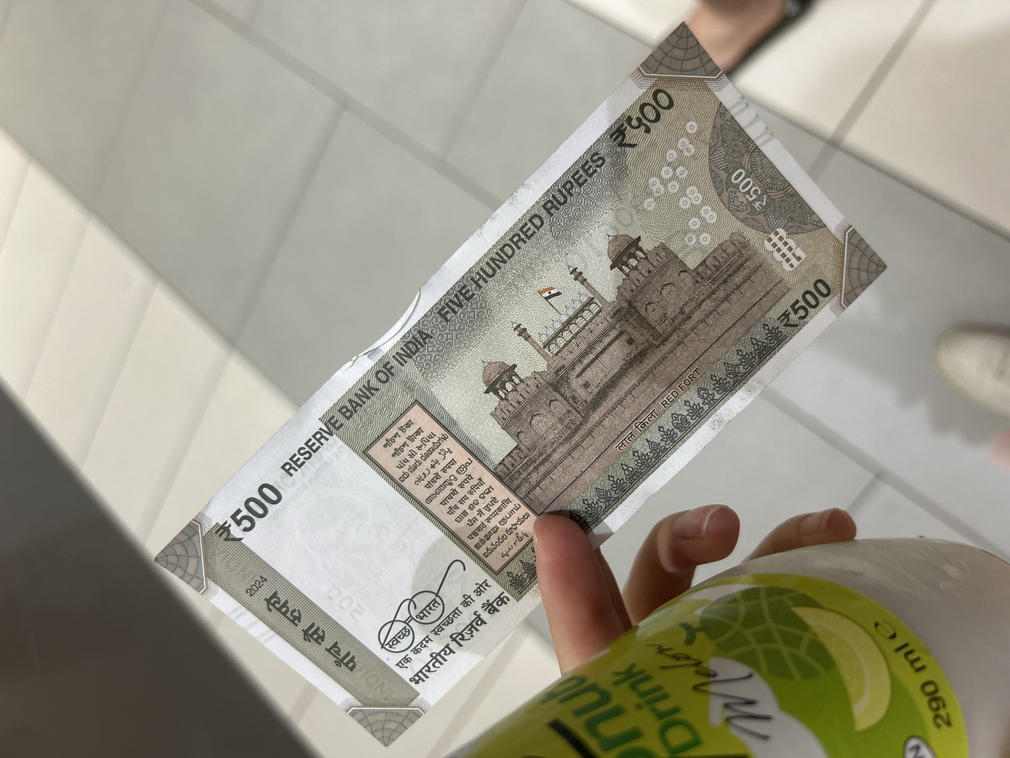 A person is holding an Indian five hundred Rupee note with their hand, also holding a 290ml Melon drink. The background appears to be a white or light colored surface, possibly tiled. The note features an image of the Red Fort. There is no indication of the location being Sofia, Bulgaria.