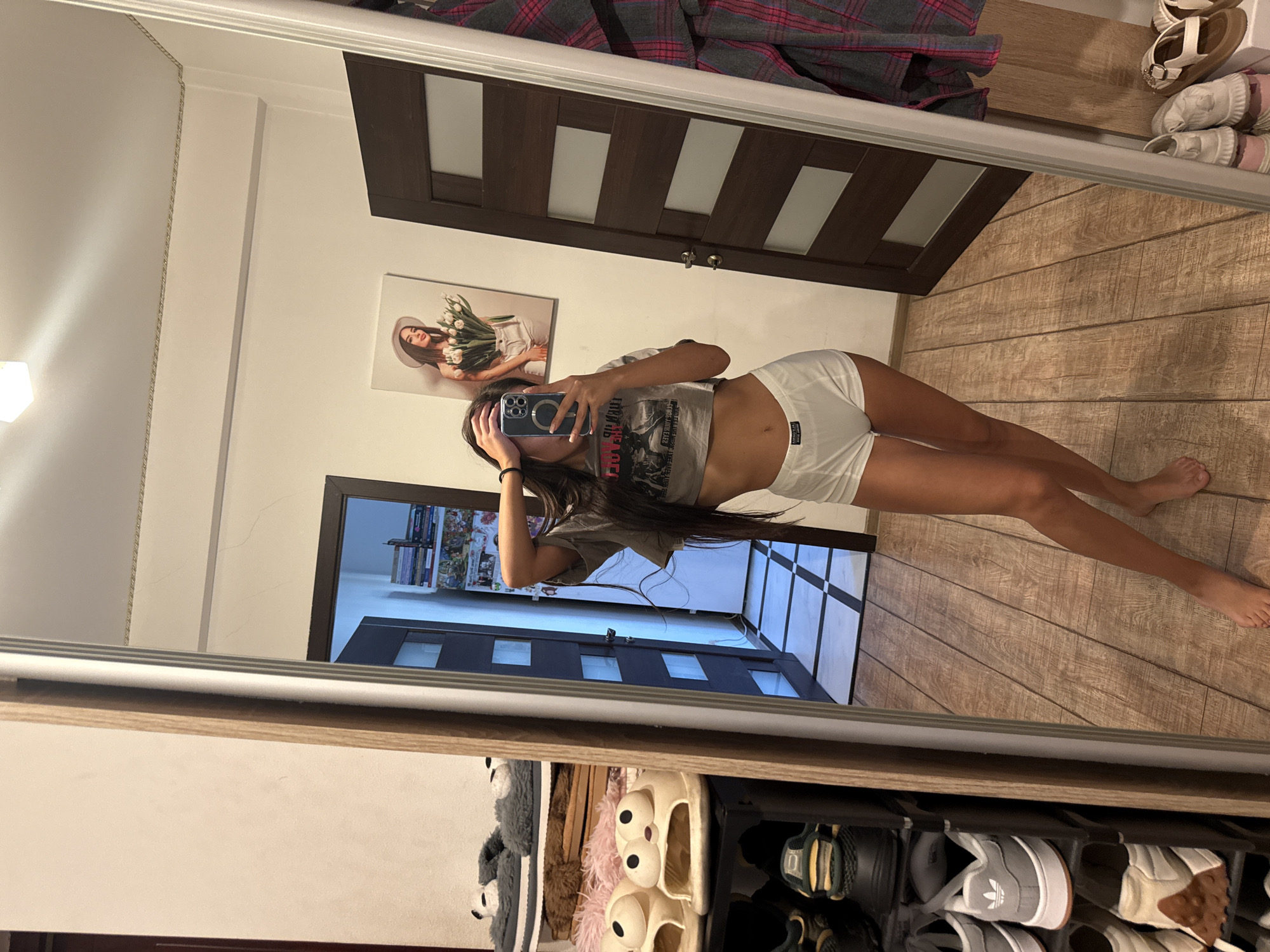 The image shows a young woman taking a mirror selfie in what appears to be a residential interior, likely in Chișinău, Moldova, based on the provided context. She is standing in front of a large mirror, wearing a grey crop top and white shorts. Her left arm is raised, holding her phone to take the picture, and her right hand is near her head.
To her left, reflected in the mirror, is a shoe rack filled with various pairs of shoes and slippers, including some fuzzy panda-themed slippers and open-toed slides with cartoonish eyes. A dark wooden door with horizontal frosted glass panels is visible behind her. On the wall, a framed picture depicts a woman in a hat holding a bouquet of flowers.
The floor is covered with wood-like tiles arranged in a diagonal pattern. Further into the background, another door is visible, and part of a hallway with checkered tiling. To the far right of the mirror, a plaid garment, possibly a jacket or blanket, is draped over a surface. The overall environment suggests a casual, domestic setting.