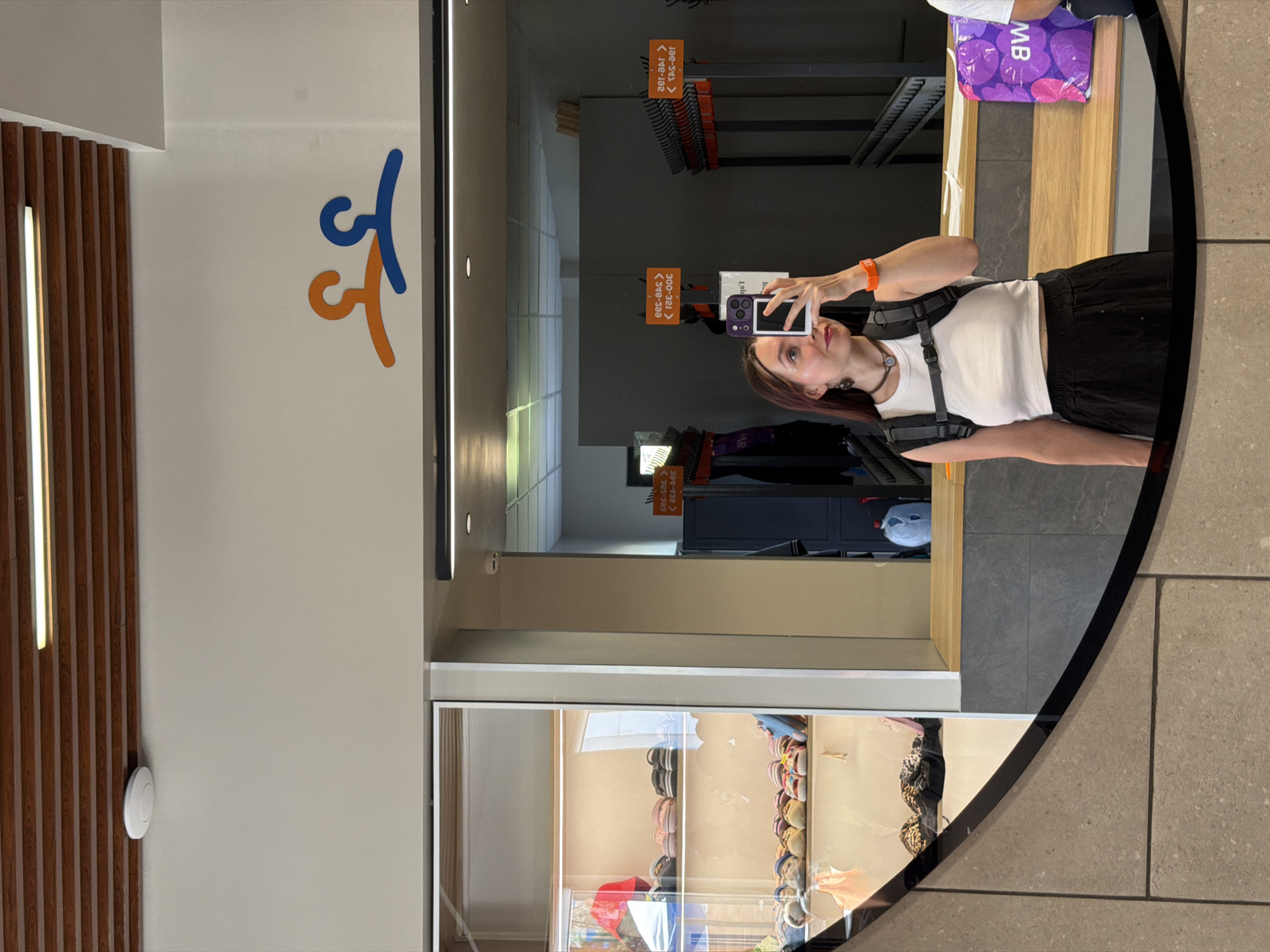 The image shows a young woman taking a selfie in a well-lit indoor space. She is wearing a white tank top, a black skirt, and a backpack. She is holding up her phone to take the picture, and is looking at her reflection in a large mirror.
The background of the image shows a shoe store display on the left, with shelves filled with various pairs of shoes. On the right side of the mirror, there are rows of empty coat hangers, suggesting a changing room or a retail area. The overall environment appears to be modern and clean. There is a sign with two colorful clothes hangers on the wall above the mirror, which appears to be a logo or decoration. The floor is tiled.
The scene is a retail store, possibly a shoe store or a clothing store, in Votkinsk, Russia. The woman appears to be a customer browsing the store, or perhaps an employee taking a break.