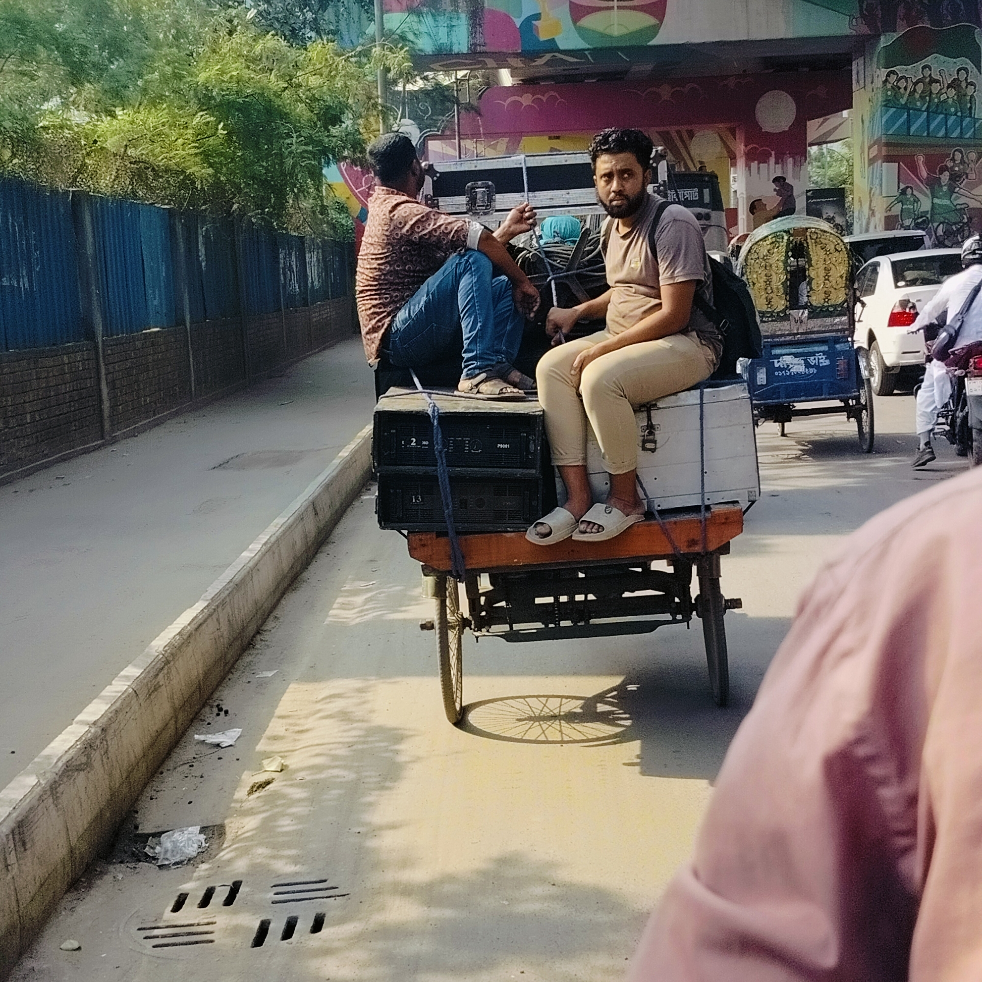Two men are riding on a three-wheeled cart in Dhaka, Bangladesh. One man is sitting on top of black storage containers, wearing a patterned shirt and blue jeans. The other man is sitting on a white container, wearing a tan t-shirt and khaki pants. The cart is being pulled down a street with a concrete curb on the left. Behind the cart, there is a blue rickshaw, a white car, and a person on a motorcycle. In the background, there is a bridge with colorful murals on the pillars. The environment appears to be a busy street in Dhaka.