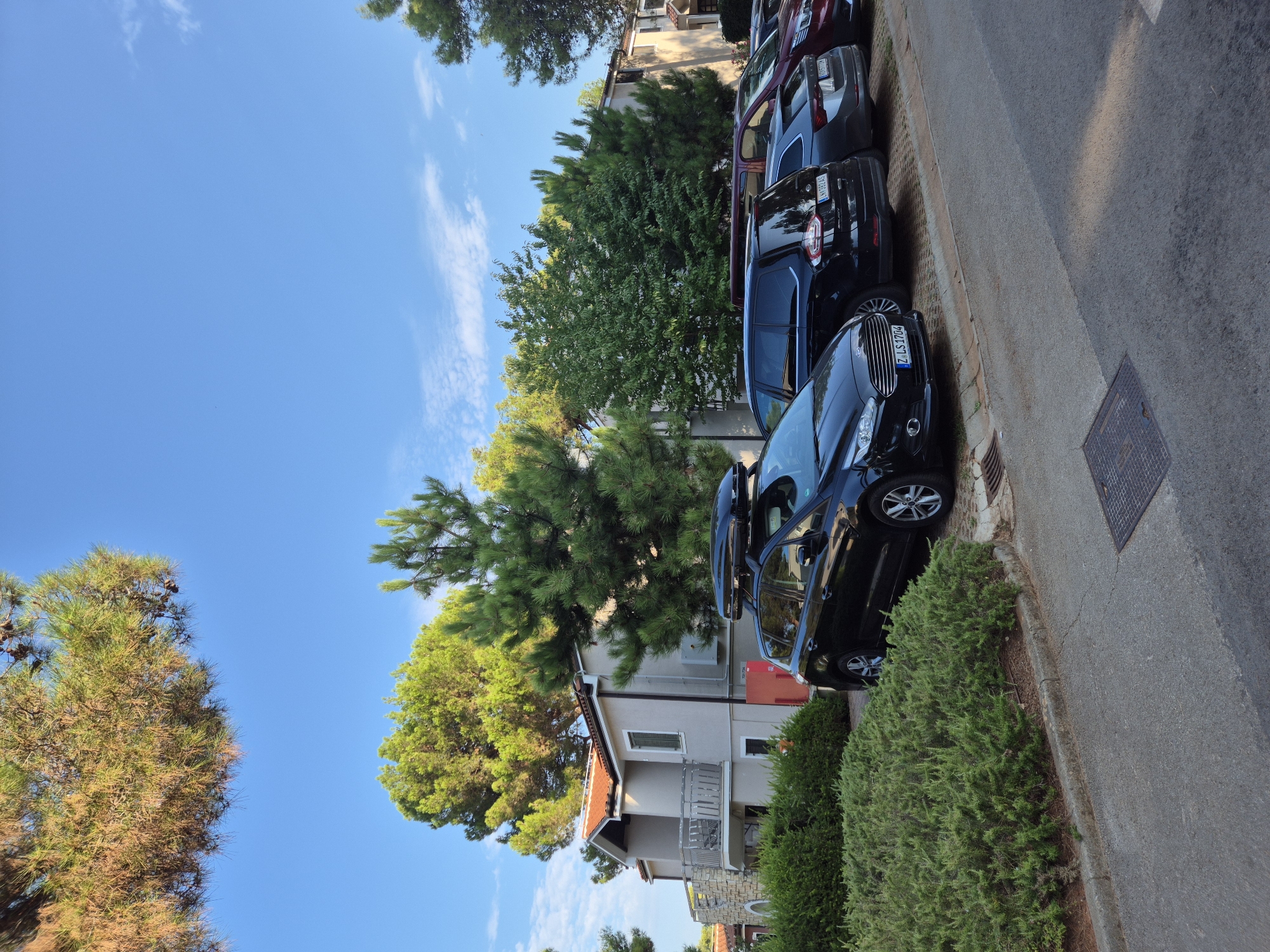 The image depicts a parking lot in front of a multi-story building in Umag, Croatia. The building is light-colored with a red-tiled roof and features balconies and stone accents. Lush green bushes and tall pine trees surround the building and the parking area. Several cars are parked in a row, including a black SUV with a roof box, a dark minivan, a grey sedan, and a red sedan. The ground is paved with asphalt, and a metal grate is visible in the foreground. The sky is clear blue with a few scattered white clouds, and the sun is casting shadows. The overall impression is a sunny day in a residential or resort area.