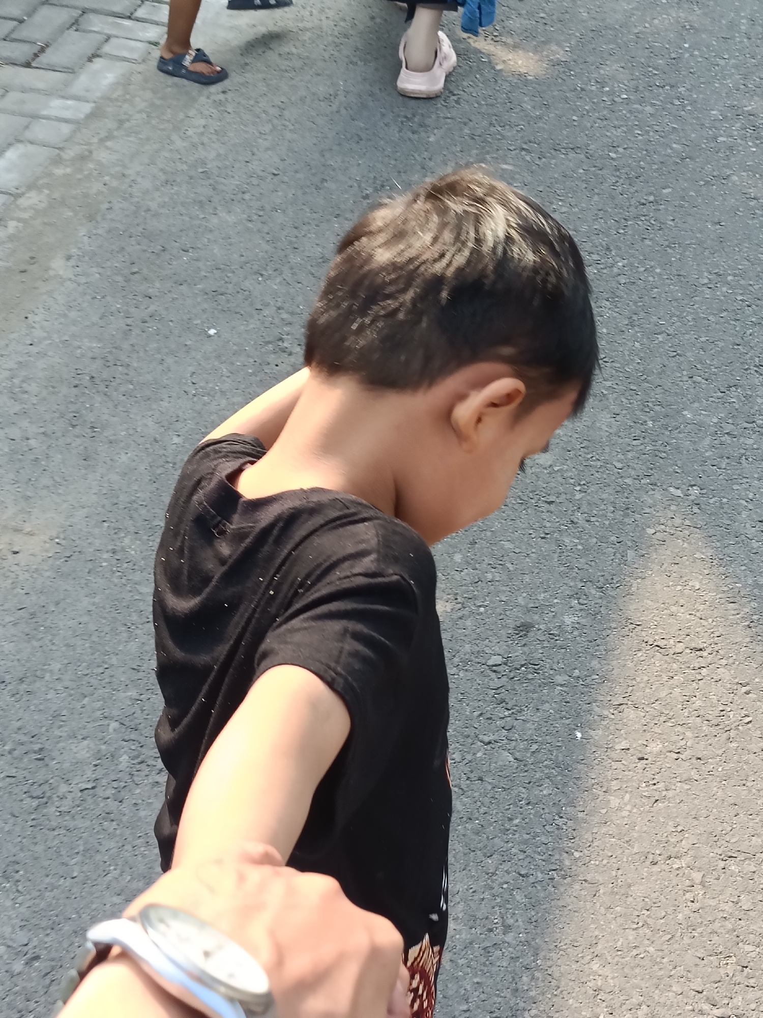 The image shows a young boy from behind, wearing a black t-shirt. He is standing on an asphalt road, with his head turned downwards and to the right. His right arm is bent and resting on the back of his neck. In the foreground, a person's arm and hand are visible, wearing a watch on the wrist. This suggests the photo is taken from the perspective of someone holding the boy's hand, possibly guiding him.
In the background, to the left of the boy, another person's legs are visible, wearing dark sandals on a paved surface. Further in the background, another person is wearing white shoes on the paved surface. The scene appears to be outdoors, with sunlight casting shadows, indicating it is daytime. The ground is asphalt, suggesting a street or a similar outdoor area. The weather appears to be sunny. No text is visible in the image. The overall impression is of a casual outing, possibly in a city environment like Kediri City, Indonesia.