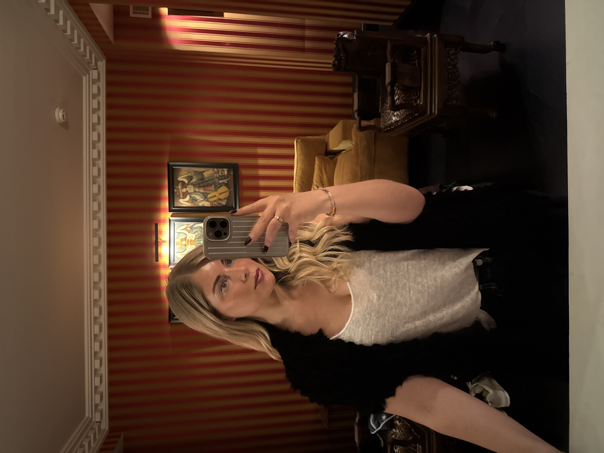 A woman is taking a selfie in what appears to be a room with distinctive interior design. The walls have red and gold striped wallpaper, and there are framed artworks visible. She is holding a smartphone, and the reflection suggests she is in a room with an ornate chair or table. The location cannot be determined to be in Paris, France, based solely on the visual content.