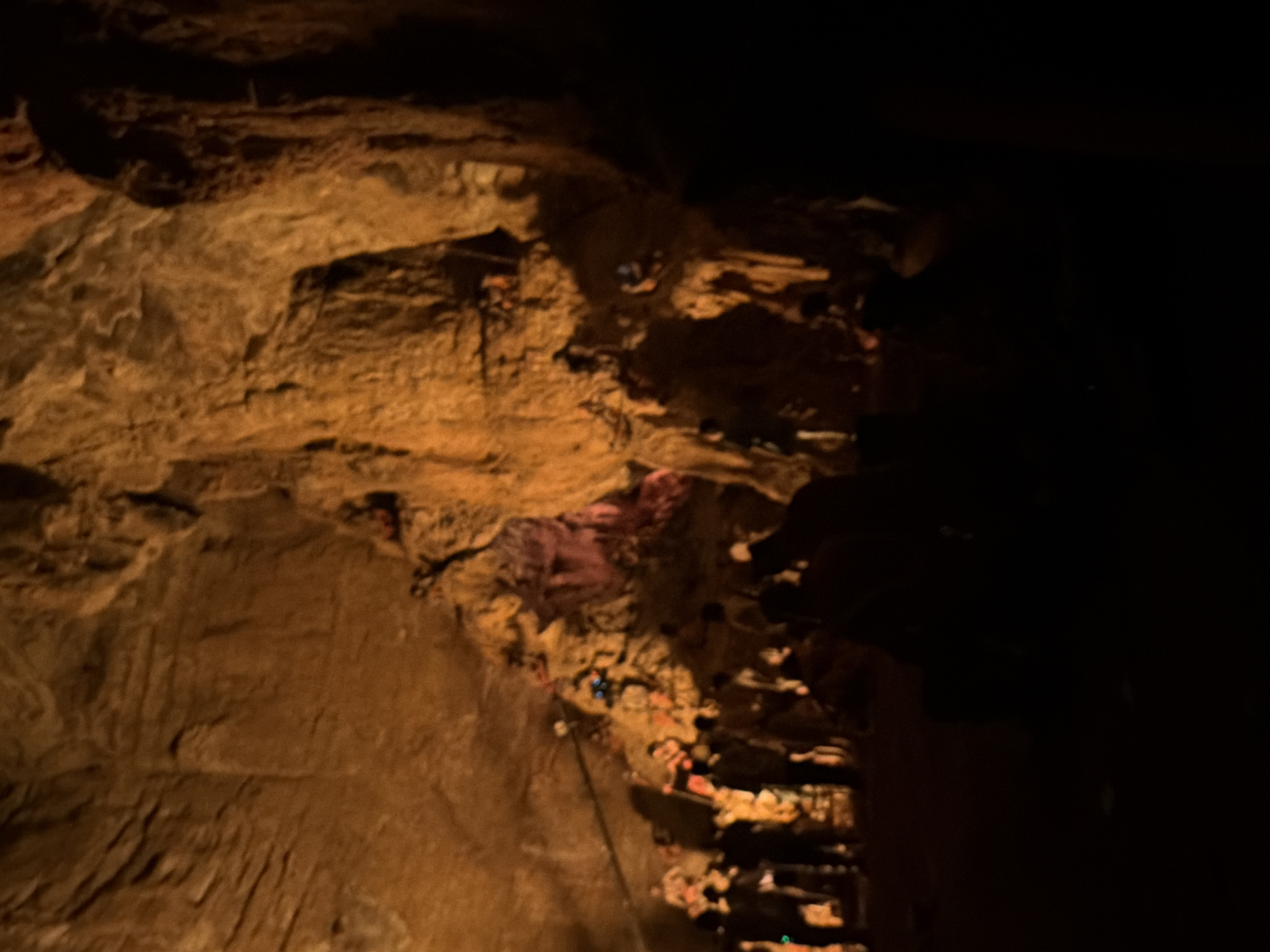The image depicts a dimly lit cave interior, likely illuminated by artificial lighting which casts a warm, orange glow on the rock formations. The cave walls are textured and uneven, with visible strata and stalactite-like formations. In the mid-ground and background, a group of people, silhouetted against the light, are gathered. They appear to be observing something within the cave. Some individuals are holding what might be flashlights or cameras. The overall impression is that of a guided tour or a gathering of explorers within a natural cavern. The scene is dark, with large portions of the foreground obscured by shadow. The location is identified as the Guttapadu Caves in Guttapadu, India.