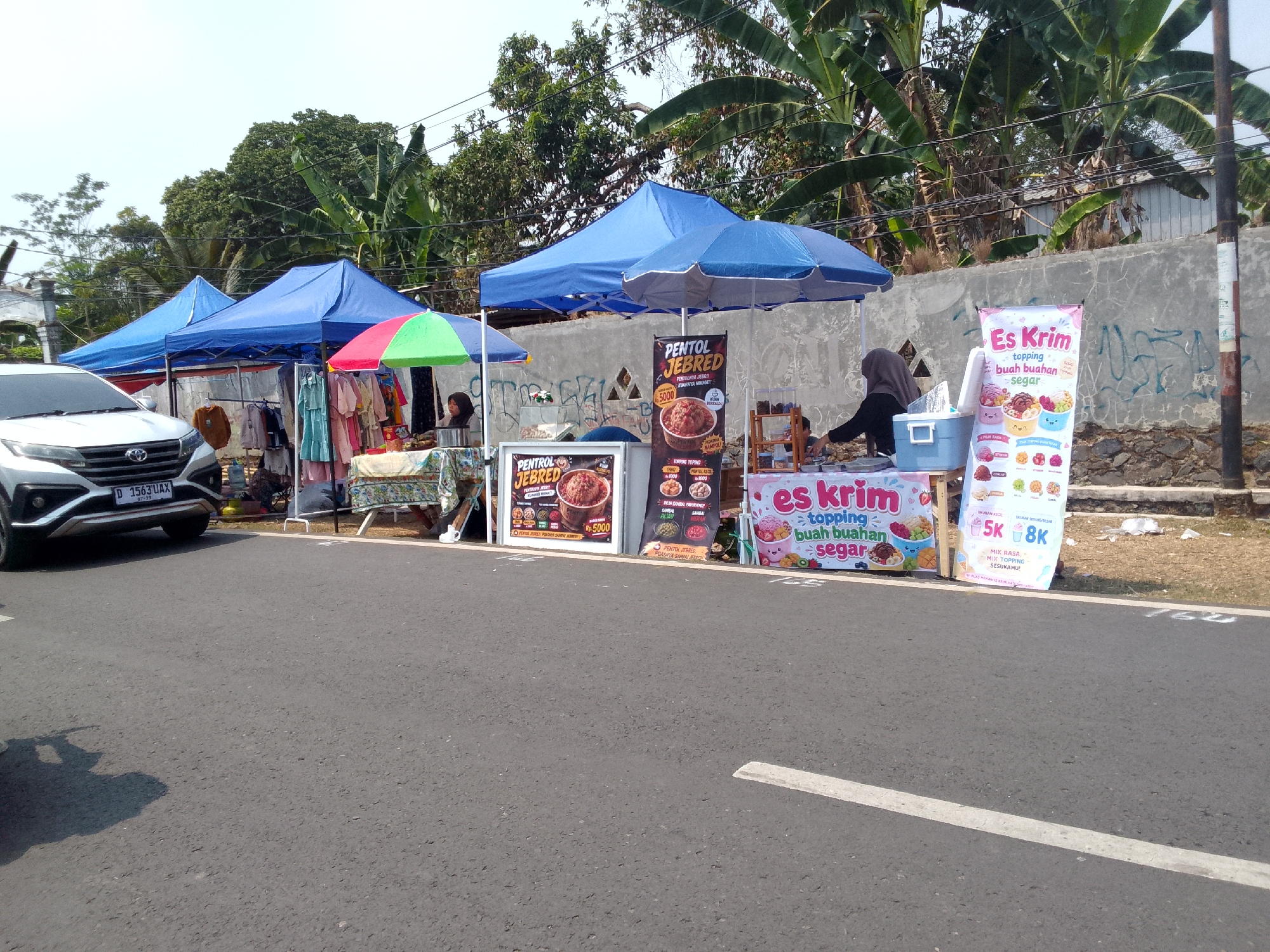 In Mandalawangi, Indonesia, on a sunny day, several food stalls are set up on the side of a paved road. A white SUV with license plate D 1563 UAX is parked to the left. Two blue tents are set up, with one displaying clothing for sale. A third, smaller tent has a red and green umbrella.
One stall features a sign advertising "PENTOL JEBRED" (spicy meatballs) with prices indicated as Rp5000. Another stall has a sign for "Es Krim" (ice cream) with various toppings such as fruit, chocolate, and sprinkles. A person in a grey hijab is seen serving customers at this ice cream stall.
The stalls are arranged near a low concrete wall, with trees, including banana trees, visible in the background. The overall scene depicts a lively street-side market.