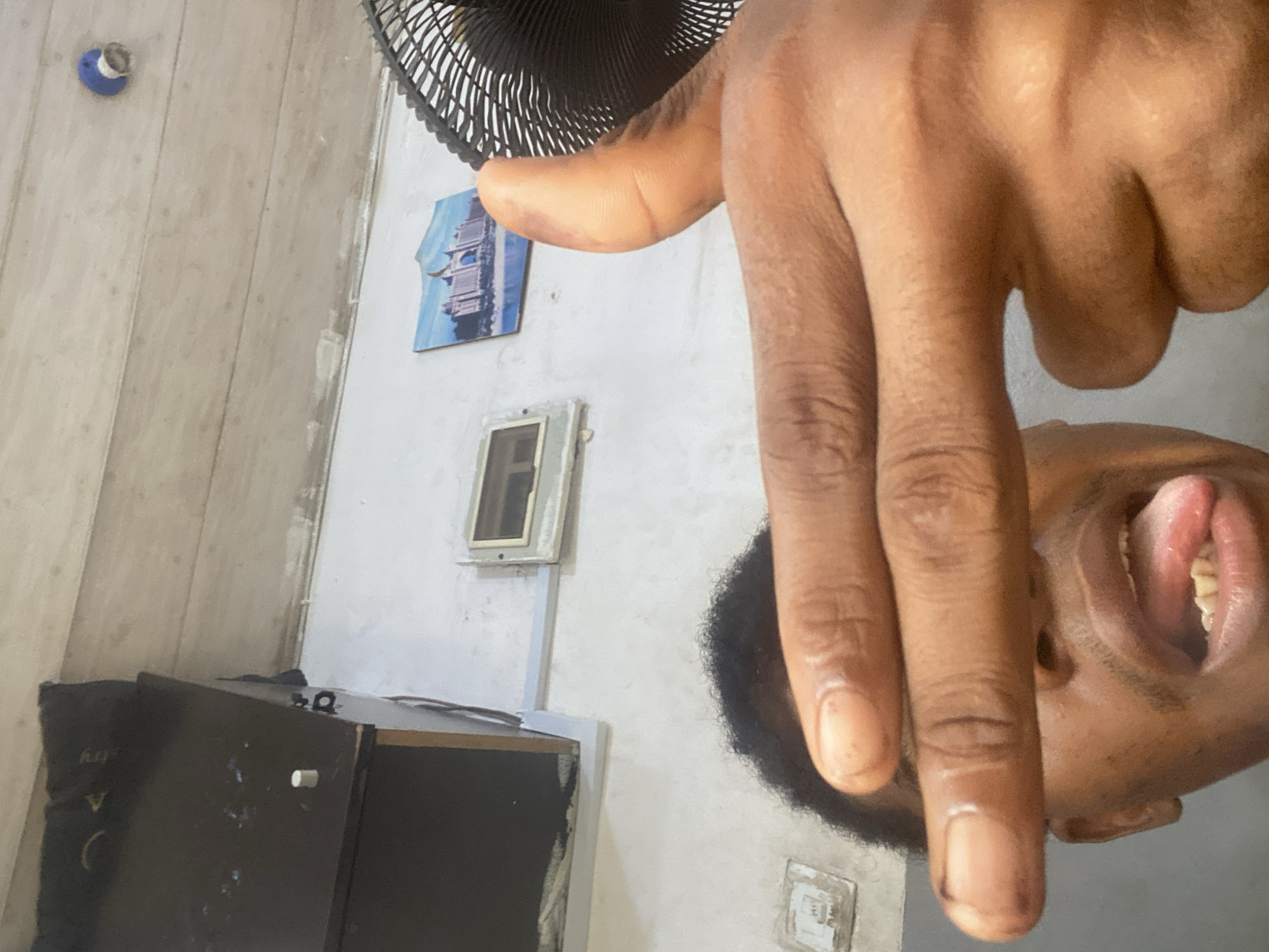 This image is captured in a portrait orientation but is rotated 90 degrees counter-clockwise, showing a close-up of a person's head and hand upside down relative to the background.
In the foreground, a person with dark skin and short, curly black hair is visible. Their mouth is open, with their tongue slightly extended, revealing their teeth, which suggests a playful or cheeky expression. A prominent hand, likely belonging to the same person, occupies a significant portion of the frame, positioned above the person's face. Part of a black electric fan grill is visible behind and to the side of the hand.
The background reveals an indoor setting with a light-colored wall, possibly white or off-white, which appears somewhat worn in areas. On the wall, there's a small blue-framed picture featuring a modern building, possibly a skyscraper like the Burj Al Arab. Below it, another rectangular fixture, resembling an empty frame or a recessed light cover, is visible. An electrical outlet or switch plate is also seen on the wall towards the lower right. To the lower left, a dark piece of furniture, possibly a cabinet or desk, is present, displaying some light-colored marks and a white cylindrical peg. Faint, unreadable text is partially visible on the furniture's surface. What appears to be a light-colored wooden planked floor or ceiling is visible in the upper left section of the image. A blue and white circular light fixture or ceiling rose is mounted in the top-left corner. The lighting is bright, suggesting the photo was taken indoors during the day or in a well-lit room.