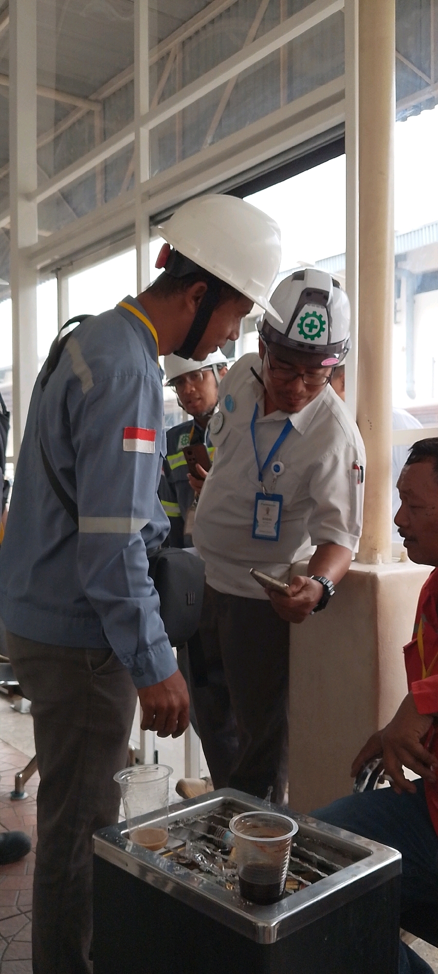 In an indoor industrial setting, likely within a factory or workshop in Depok, Indonesia, four men are gathered around a metal receptacle that appears to be an ashtray. Two of the men are wearing hard hats and safety vests over their work attire, indicating a work environment. One man, in the foreground, is wearing a blue uniform with a red and white Indonesian flag patch on his left sleeve. He is looking down and to his right, seemingly engaged in conversation or observation with the man beside him. The second man in a hard hat, in the center of the frame, is wearing a light-colored shirt and holding a smartphone, also looking down and to his right. A third man, partially visible in the background, is also wearing a hard hat and safety vest. The fourth man, on the far right, is wearing a red shirt and appears to be looking at the men in the center. The ashtray contains cigarette butts and two plastic cups, one with a light-colored liquid and the other with a dark liquid. The environment features exposed beams and a window in the background, suggesting a covered structure. The lighting is ambient, typical of an indoor industrial space.