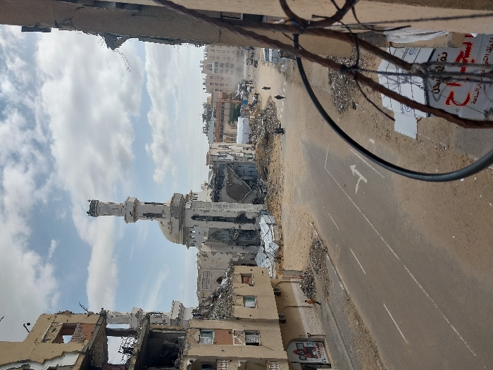 This image captures a wide view of a severely damaged urban landscape in Khan Yunis, Palestinian Territory, under a partly cloudy sky. At the center, the Grand Mosque of Khan Yunis stands with its dome and minaret partially intact but showing extensive structural damage, including visible scarring and large sections of its complex reduced to rubble. To the left, a multi-story building is significantly collapsed, with exposed rebar and debris spilling onto the adjacent sidewalk. Another damaged structure, partially out of frame, is visible on the far right, featuring exposed elements and a partial sign with Arabic text.
A paved road with faded white lane markings and a right-turn arrow runs through the foreground and midground, appearing dusty and strewn with sand and small debris. Large piles of rubble, twisted metal, and shattered building materials are scattered across the scene, particularly around the damaged structures and in the open areas.
On the left side of the road, near a building entrance, one individual in a yellow top and dark pants walks towards the left. Further down the road, in the distance, at least four other individuals are visible walking. A black cable and rusted rebar frame the lower right foreground, suggesting the image was taken from an elevated vantage point within a damaged building. The overall environment depicts widespread destruction and a dusty atmosphere.