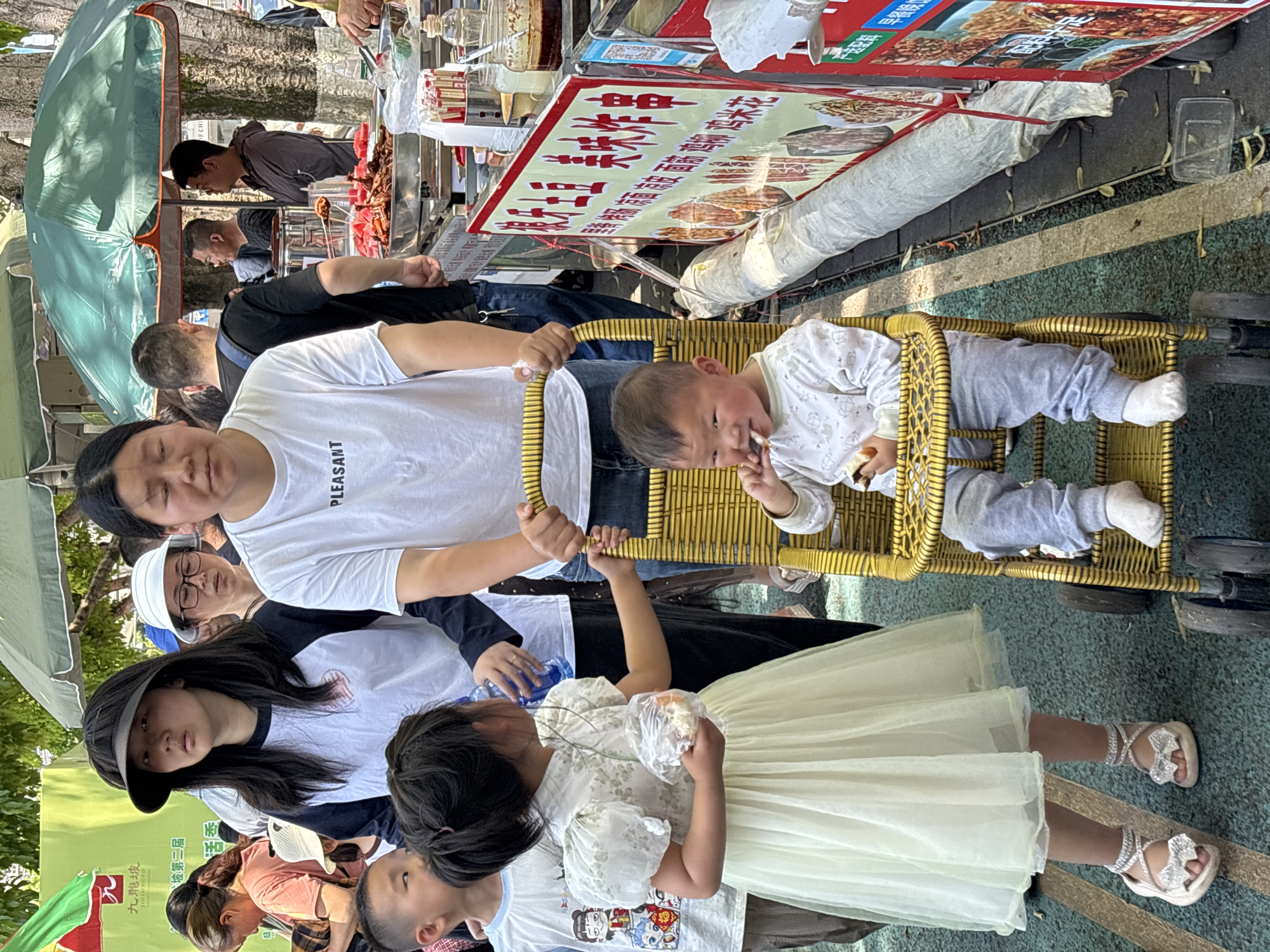A group of people, including adults and children, are gathered in what appears to be a public outdoor space. A baby is sitting in a small, gold-colored woven chair and appears to be eating. An adult in a white t-shirt is holding the chair. To the right of the child is a food stand, with a large advertisement in Chinese script displayed, with some individuals seemingly interested in the food offerings. Another child is holding a piece of food. The environment suggests an urban setting, possibly in the Jiulongpo District, China, based on the text visible on a sign.