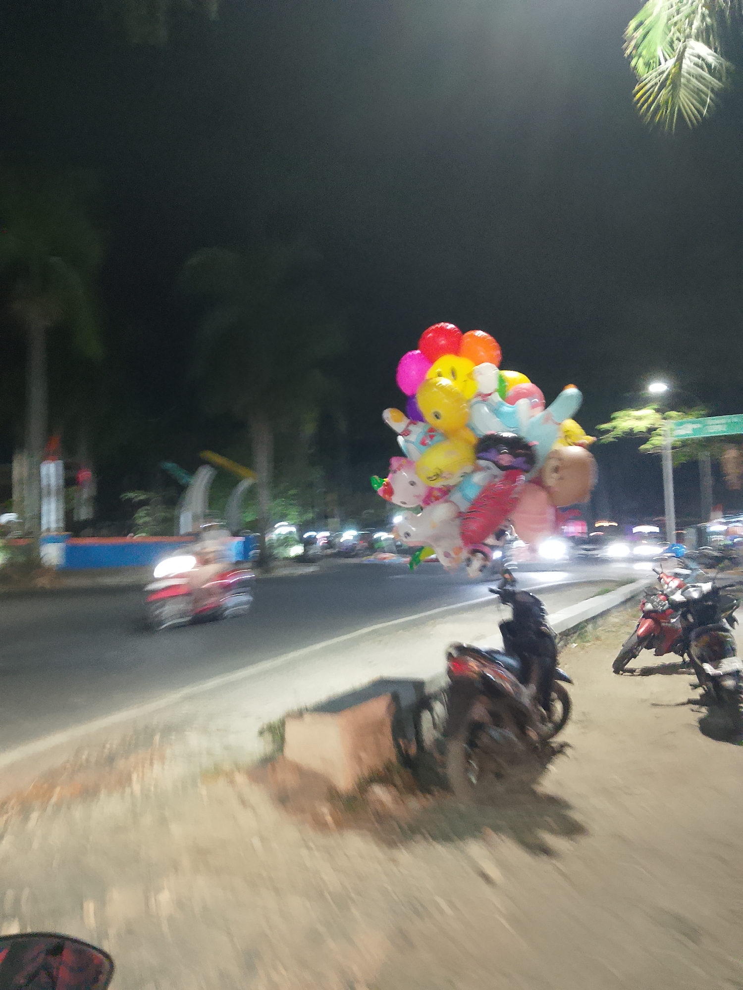 A bunch of colorful balloons shaped like cartoon characters is being sold at night on a street in Balun, Indonesia. The balloons are being held up by a seller who is not fully visible in the frame. The street is lit by streetlights and the headlights of passing vehicles. There are several motorcycles parked on the side of the road. The scene is blurry due to low light conditions and motion.