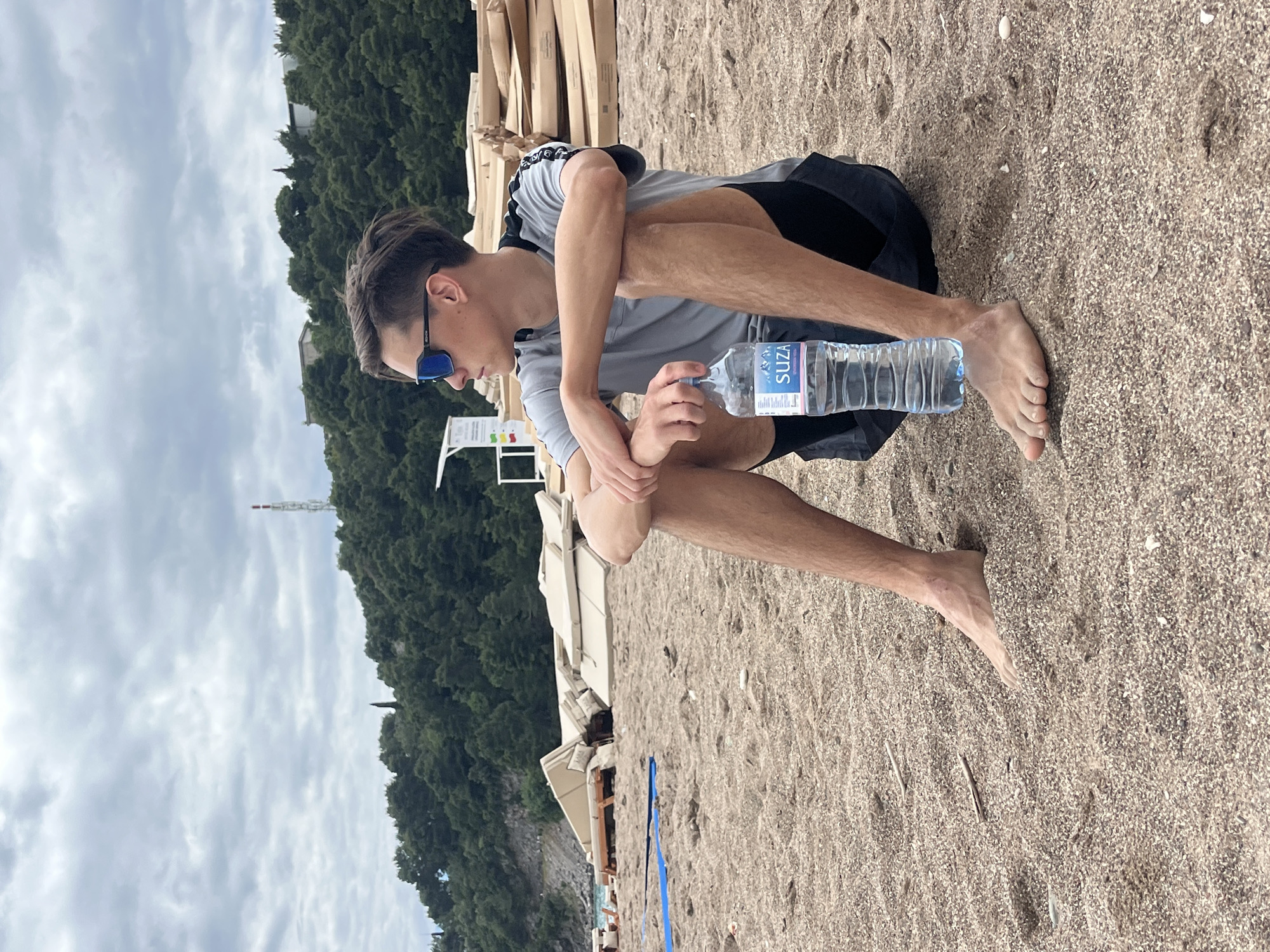 A young man sits cross-legged on a sandy beach in Boreti, Montenegro. He wears a grey t-shirt, black shorts, and blue sunglasses. He is holding a plastic bottle of SUZA water. Behind him are beach chairs and a line of trees. In the background, a grey, cloudy sky is visible. A tall, thin, white tower is seen rising from the trees.