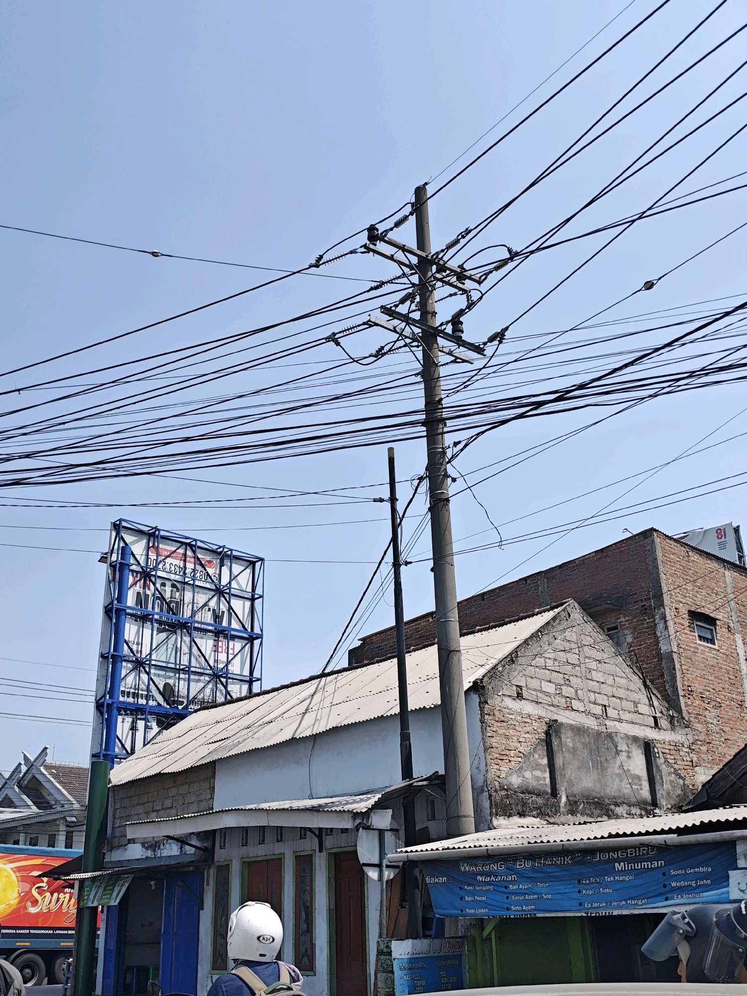 The image captures a street scene in Gampengrejo, Indonesia, under a clear blue sky. A prominent utility pole with a dense network of power lines dominates the center-right of the frame. In the background, a large blue and white billboard stands tall, its text partially obscured by the utility pole and the angled perspective. To the right of the billboard, a weathered brick building rises, its upper stories visible. In the foreground, several low-rise buildings with corrugated metal roofs line the street. One building displays a blue banner with text that appears to be a menu, listing "Warung Bu Hanik" and various food and drink options. A person wearing a white helmet and backpack is partially visible in the lower right corner, facing away from the camera, suggesting they are on a motorcycle. To the left of the buildings, the side of a red truck with yellow lettering, possibly advertising "Swirl," is visible. The overall scene depicts a typical urban street with infrastructure, commercial activity, and pedestrian presence.