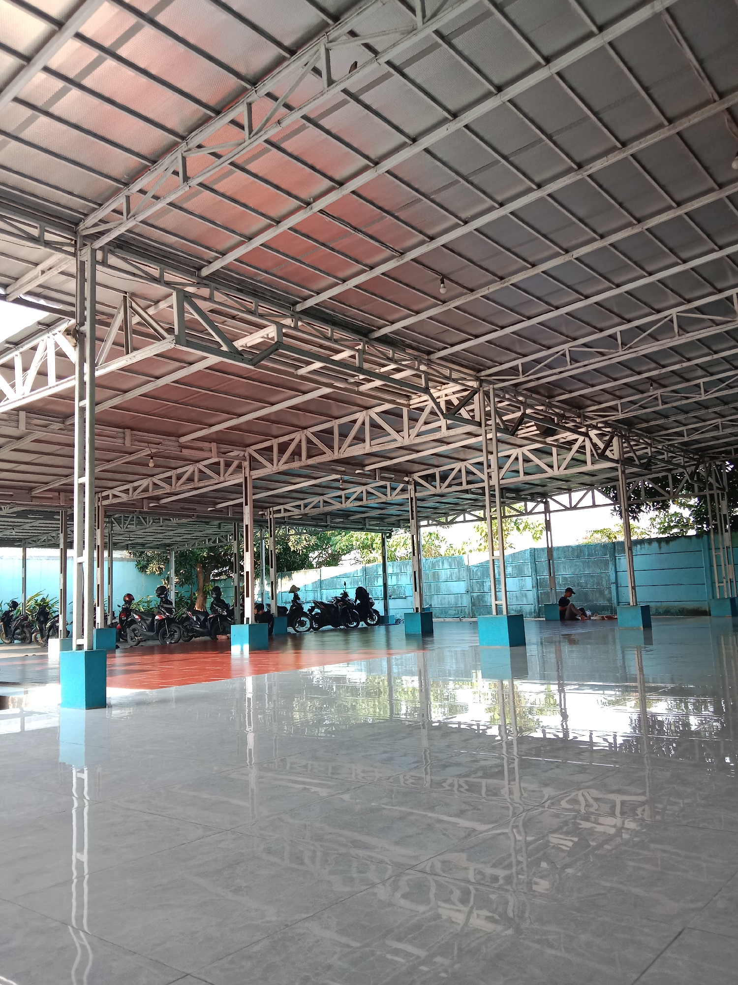 The image depicts an open-air covered area, likely a parking lot or a gathering space, with a corrugated metal roof supported by a steel frame. The floor is tiled and highly reflective, showing clear reflections of the structure and surrounding trees. Parked motorcycles are visible in the background, and a few individuals are present, some seated on the floor. The scene appears to be in Semarang, Indonesia, as suggested by the presence of typical architecture and vegetation. The overall atmosphere is one of an ordinary day in a public or semi-public space.
