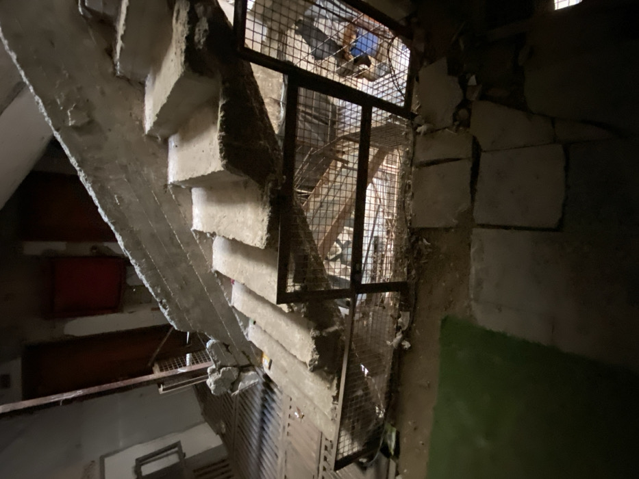 The image displays a damaged interior stairwell within a building in Khan Yunis, Palestinian Territory. A concrete staircase, extending from the upper right to the lower left, shows considerable structural damage, including cracked and broken steps, exposed aggregate, and sections of missing material. A dark, grid-patterned metal railing with a brown frame runs along the edge of the stairwell. Below and around the stairs, the environment is cluttered with debris, consisting of broken concrete fragments, dirt, and miscellaneous refuse. In the upper left background, two dark red doors are visible against a light-colored wall, alongside a window frame and a vertical metal pipe. The lower foreground presents an uneven ground surface, composed of several fractured concrete slabs and bare earth, with a small, flat patch of green material in the bottom left corner. The scene is dimly lit, and no individuals or discernible activities are present.