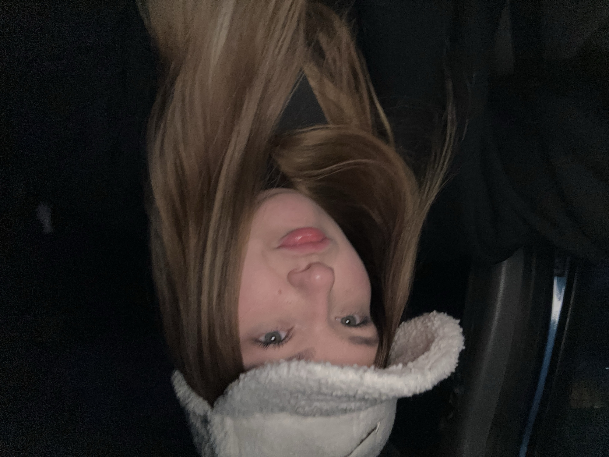 A young person with light brown hair and a fluffy white hat is indoors, possibly in a car, given the door frame visible to the right. The background is dark. The individual is facing the camera. There is no way to determine the location is in Chaikovsky, Russia.