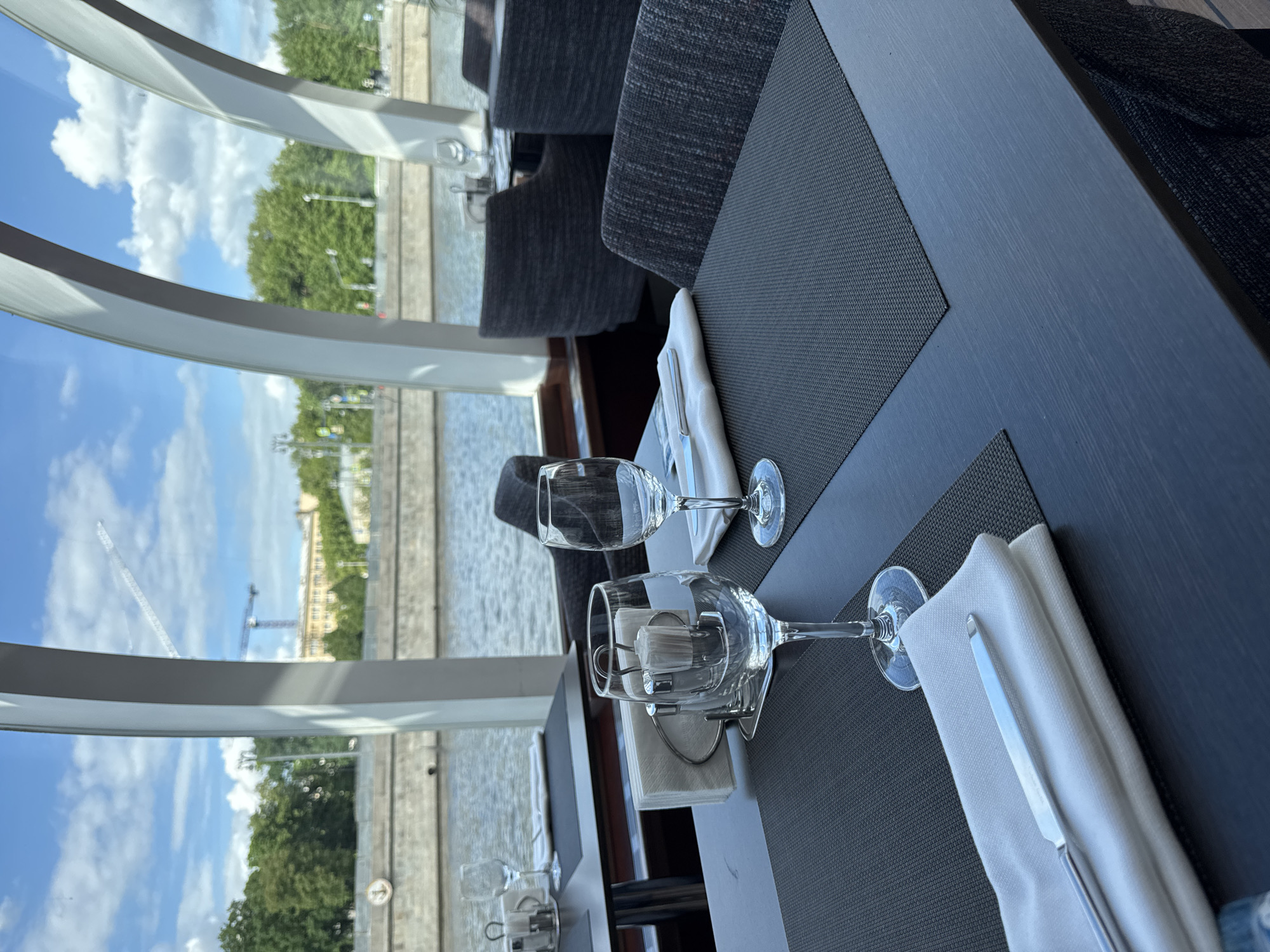 The image captures a table setting within a restaurant or dining establishment that offers a view of the Moscow cityscape in Russia. The table is set with two wine glasses, napkins, and cutlery on placemats. The interior lighting is subdued, emphasizing the view outside the window. The window showcases a river with embankments, trees, and architectural structures typical of Moscow. Notably, cranes are visible in the distance, indicating ongoing construction or development in the area. The scene suggests a fine dining experience with a scenic backdrop.