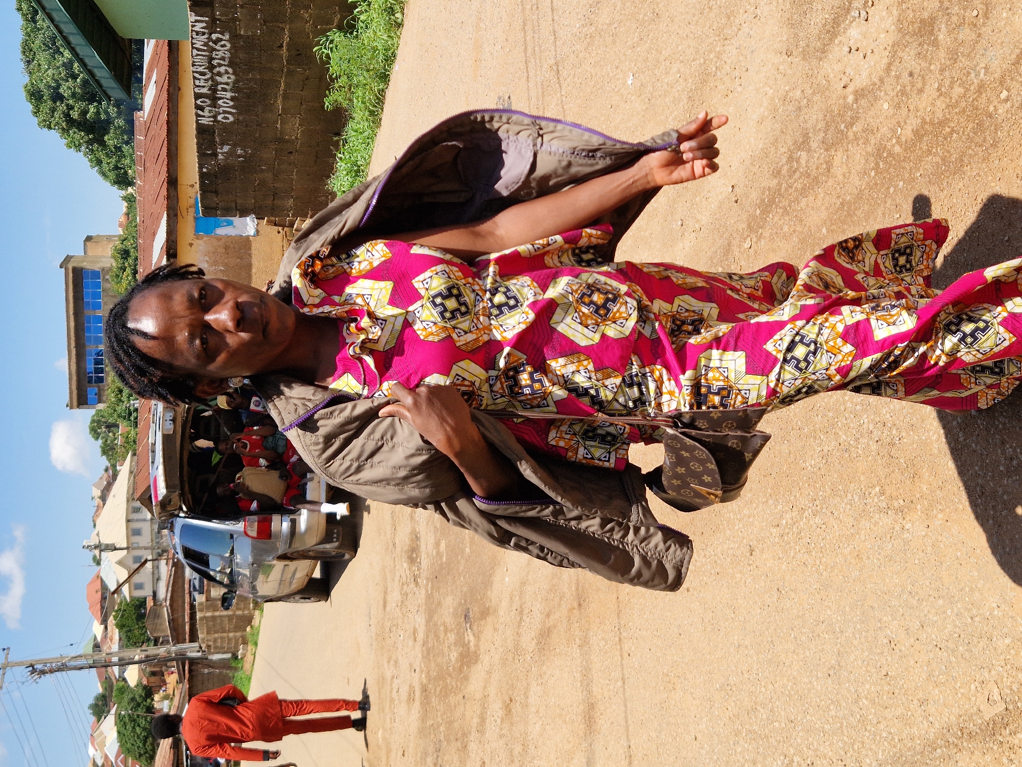 An African woman wearing a vibrant pink dress with a geometric pattern and a brown jacket walks towards the camera on a dirt road in Kabong, Nigeria. She carries a brown designer handbag on her left shoulder. Her hair is styled in braids, and she is wearing earrings. In the background, a silver car is parked with its trunk open, containing several people. A man in a red outfit walks away from the camera on the left side of the road. Buildings with corrugated iron roofs and some with blue-tinted windows are visible in the distance. A brick wall with writing on it, including "NGO RECRUITMENT" and a phone number, is present on the right side of the scene. The sky is blue with scattered clouds, and greenery is visible on the right side of the road. The scene appears to be a residential area within the city.