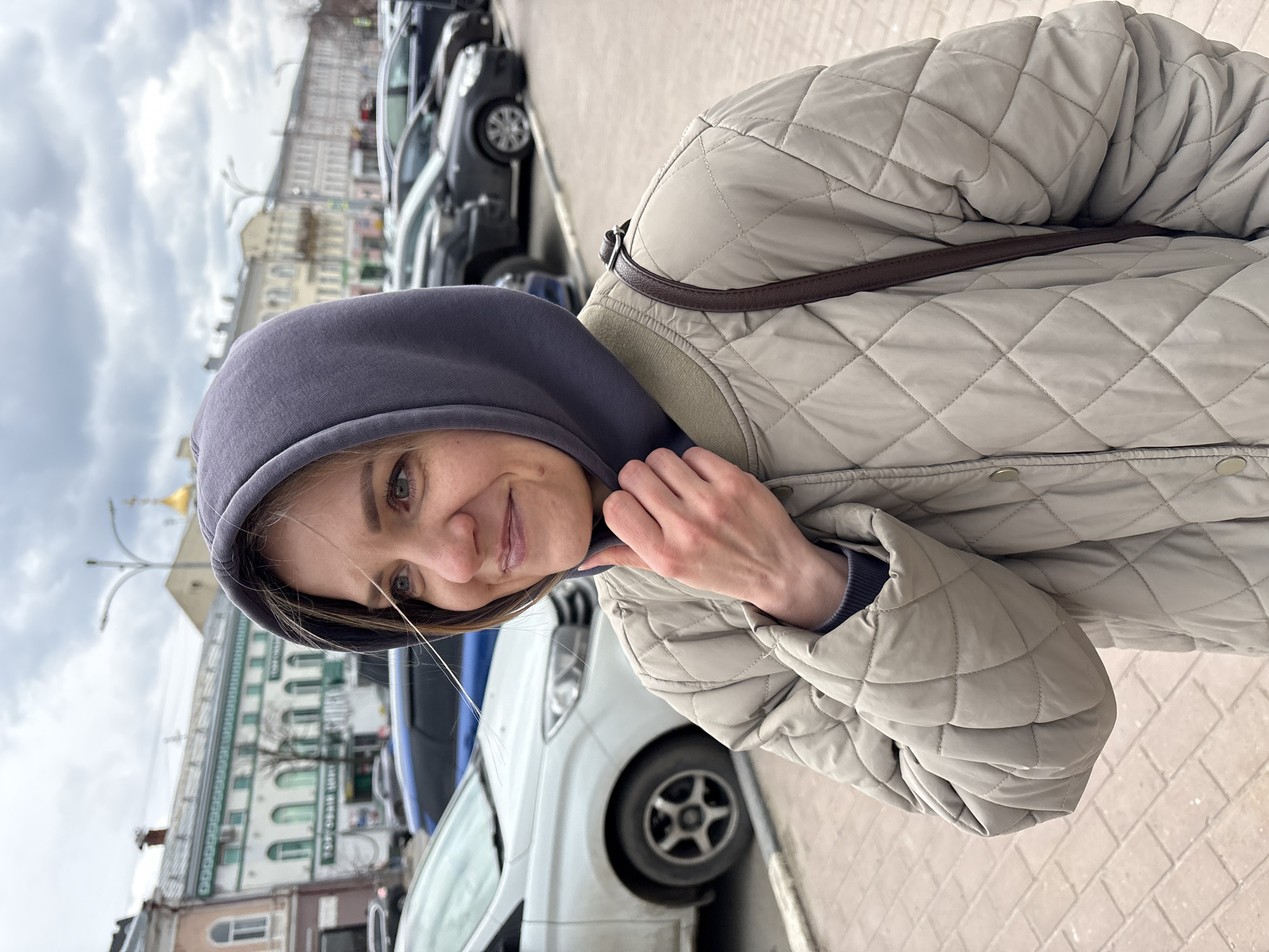 This portrait captures a young woman outdoors on a cloudy day in Ulyanovsk, Russia. She is positioned in the foreground, looking towards the camera with a gentle, pleasant smile. She wears a dark grey hooded sweatshirt under a light beige quilted jacket, with a dark brown strap (possibly from a bag) visible on her shoulder. Her right hand is lightly placed near her hood.
The background reveals an urban street scene. A line of parked cars, including a white and a black vehicle, runs alongside a paved sidewalk. Behind the cars, several multi-story buildings are visible, some with distinctive architectural features like green window frames. In the distance, a golden dome, likely belonging to a church or cathedral, stands out against the overcast sky, which is filled with grey clouds. The overall impression is of a casual moment in a city environment during daytime. No discernible text is clearly visible.