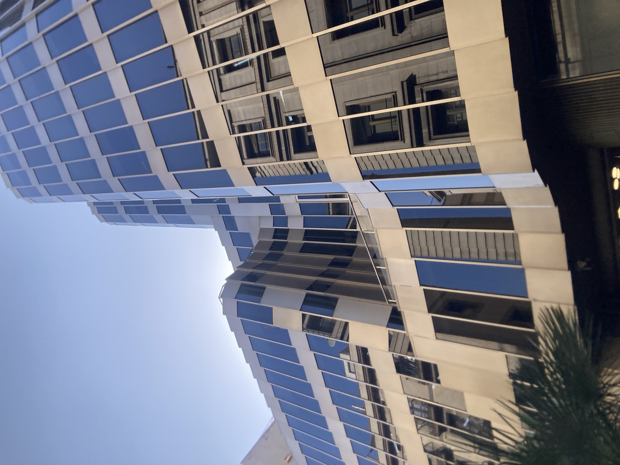 This is a low-angle, full shot of the facade of a modern building in Barcelona, Spain, with a clear blue sky above. The building features a stepped, angular design with extensive use of glass windows and metallic siding. The windows are rectangular and tinted dark blue, reflecting the sky and surrounding architecture. The metallic siding appears to be a light beige or champagne color, with horizontal bands accentuating the structure.
The reflection in the windows reveals an older, classical building with stone facades, visible window frames, and architectural details. This juxtaposition suggests the modern building is situated in a part of the city with a mix of architectural styles.
In the lower left corner, a portion of a dark green, leafy palm frond is visible, suggesting the presence of greenery or landscaping in front of the building. The lighting indicates it is daytime, likely morning or afternoon, with sunlight casting subtle shadows and highlighting the building's textures. No people are visible in the frame. The overall impression is one of urban architecture, showcasing a contrast between contemporary design and historical context.
