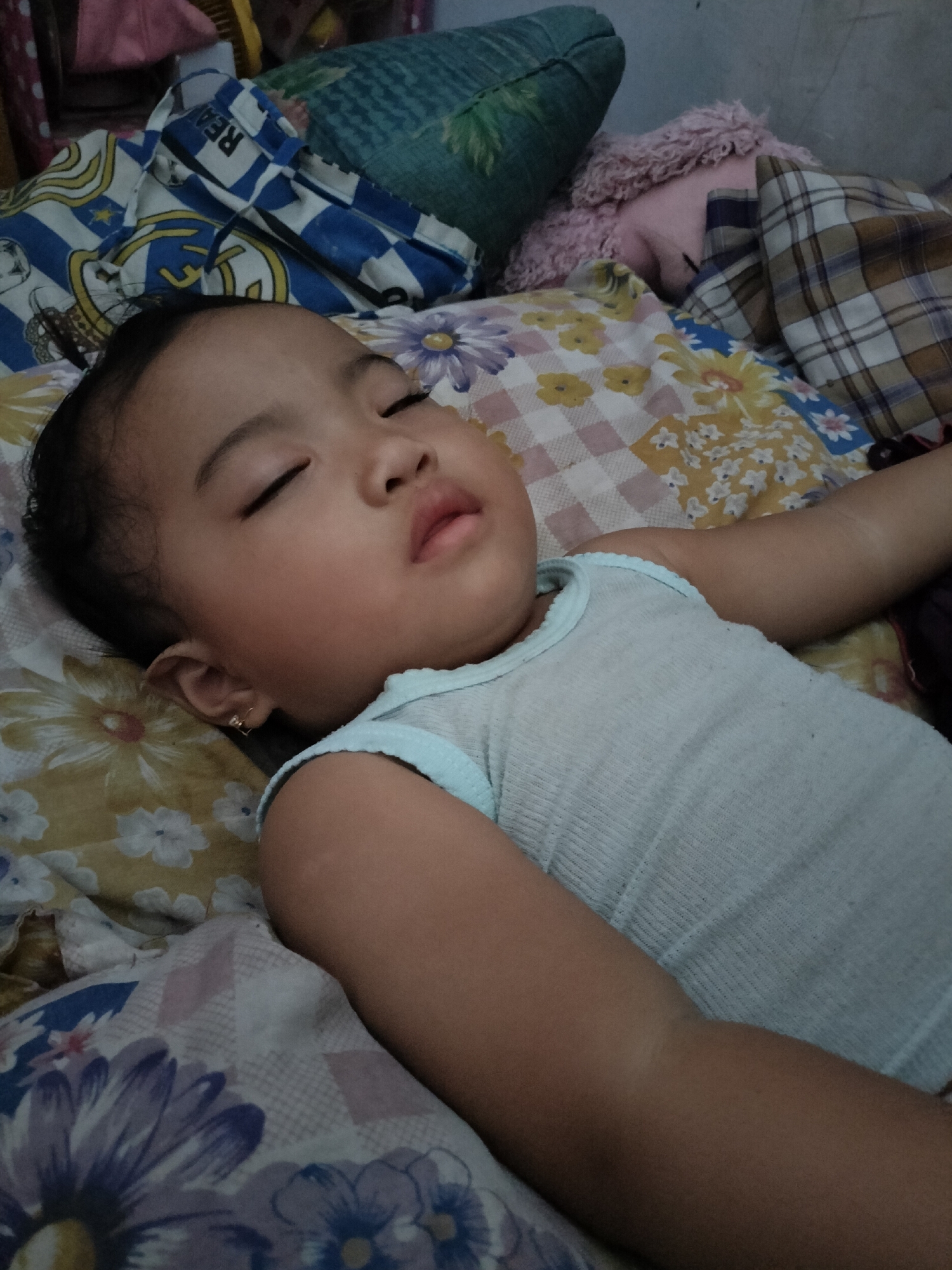 A young child is sleeping on a bed covered with floral-patterned sheets and pillows. The child is wearing a light blue tank top. The room appears to be a bedroom in a residential setting. The specific location of Leces, Indonesia is not ascertainable from the visual information provided.
