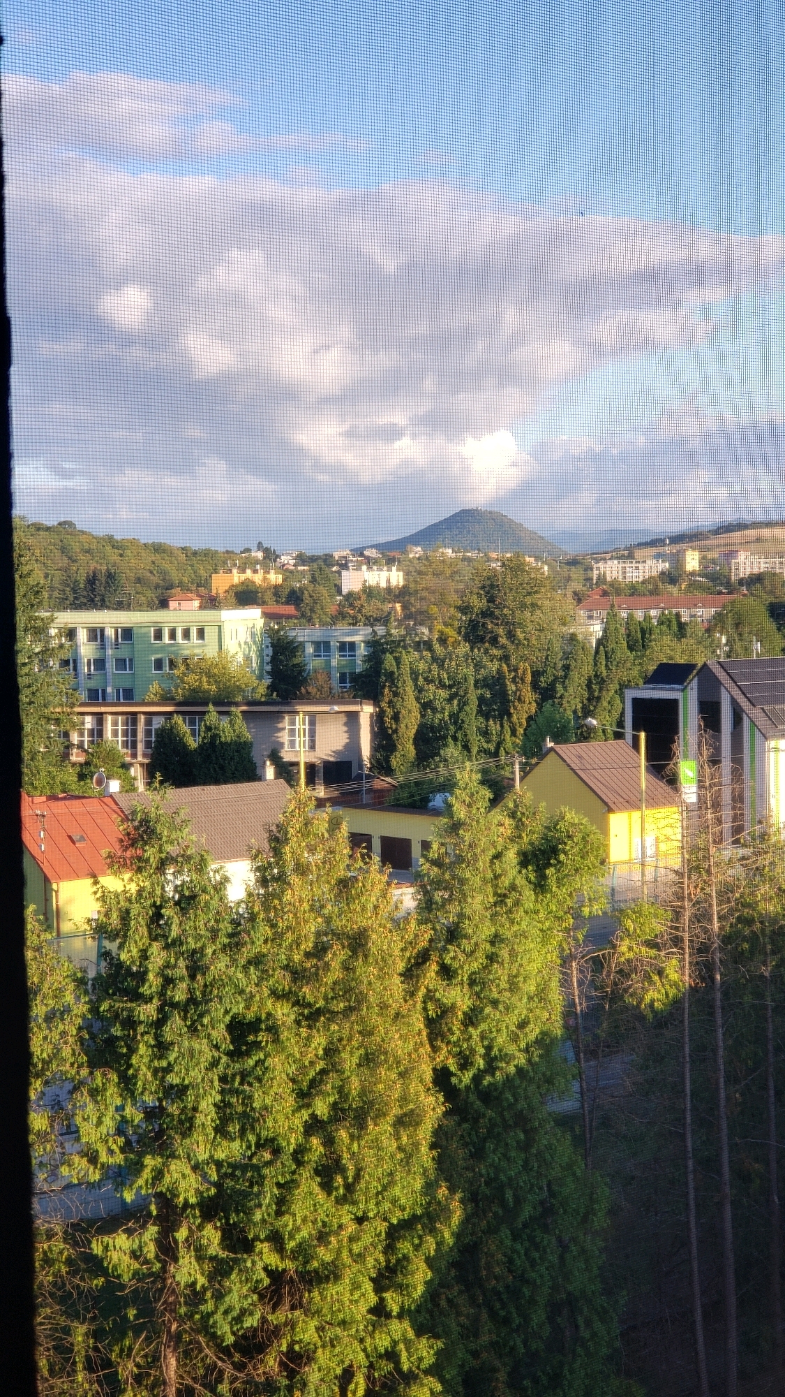 The image depicts a panoramic view of a suburban area in Prešov, Slovakia, seen through a window screen. The foreground is dominated by lush green trees, with sunlight illuminating their foliage. In the mid-ground, various residential buildings are visible. These include a long, light green apartment building with multiple stories and many windows, a grey building with darker window frames, and a smaller yellow building with a red-tiled roof. Further in the background, a conical, tree-covered hill rises majestically, with a town or city spread across its slopes and in the valley below. The sky above is a clear blue, punctuated by soft, white clouds. The overall impression is of a tranquil, sun-drenched afternoon in a well-vegetated urban fringe.