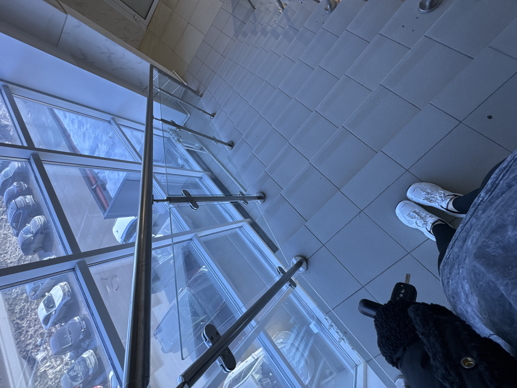 The image depicts a high-angle view from an indoor landing in the city of Chaikovsky, Russia, looking down a tiled staircase and through a large window wall to an outdoor parking area. In the immediate foreground, a person's lower torso, legs, and feet are visible. The individual wears dark leggings, white athletic sneakers, and holds a black car key fob with a metal key in a gloved left hand, adorned with a dark, textured sleeve featuring a brass-colored snap button. They stand on a light grey tiled floor.
A modern staircase, also composed of light grey tiles with integrated anti-slip ridges on each step, descends diagonally to the lower right. The staircase is bordered on the left by a clear glass and stainless steel railing system, secured by multiple vertical posts. Through the expansive window wall, which features a light-colored frame, an exterior parking lot is visible. The ground outside is partially covered with snow or slush. Several vehicles are parked, including multiple dark-colored sedans, an SUV, and a lighter-colored sedan. In the lower left background, behind a glass panel of the railing, a white radiator is partially visible. To the right of the staircase, a light-colored wall with faint, irregular grey patterning is discernible.