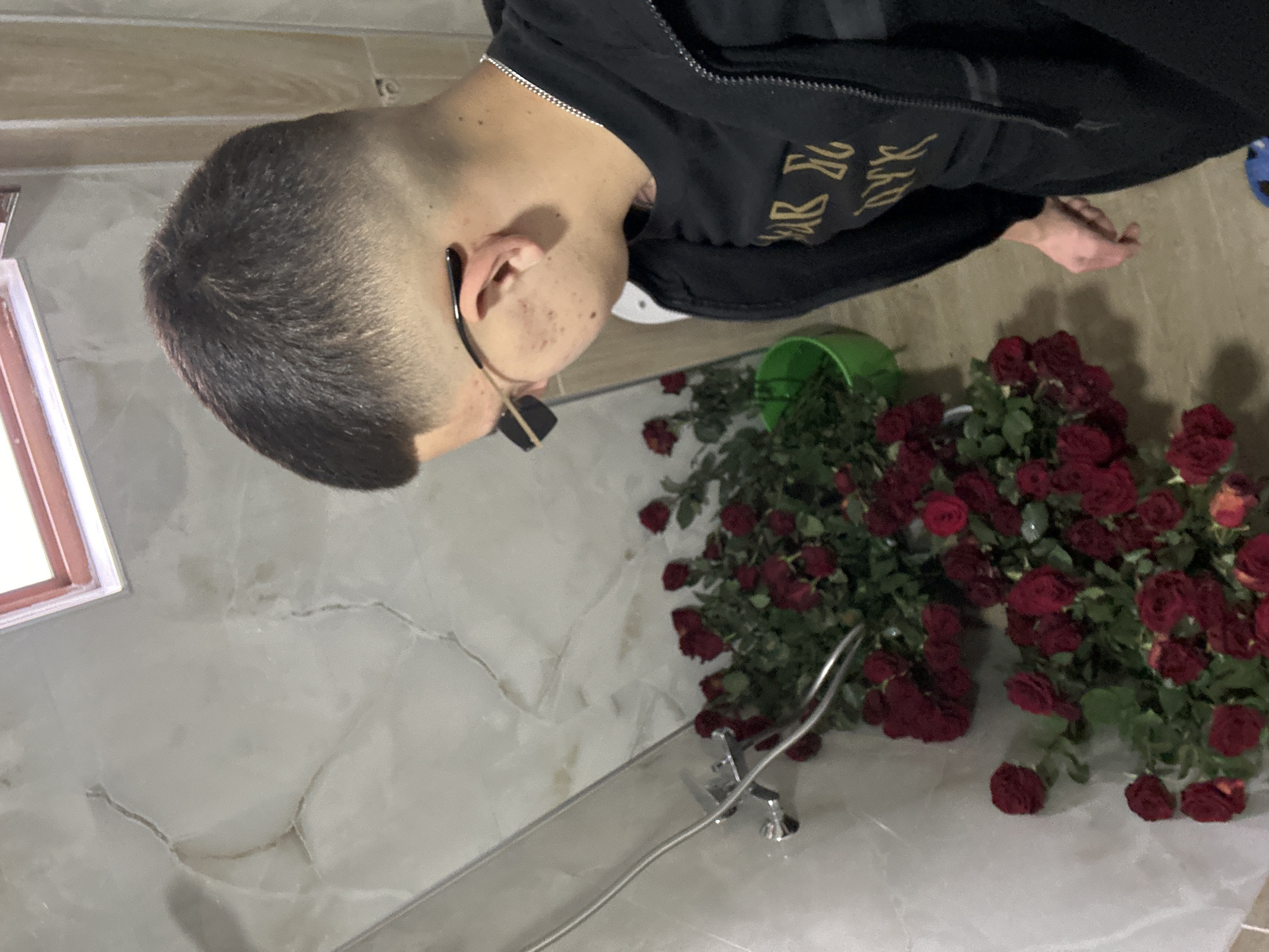 A man with short dark hair and glasses stands next to a bathtub in a bathroom. He is wearing a black hooded jacket with writing on it. A green bucket sits near the bathtub, and numerous red roses are scattered around the bucket and the tub. The bathroom has a light-colored marble floor and wall. The scene may be in Zlatograd, Bulgaria.